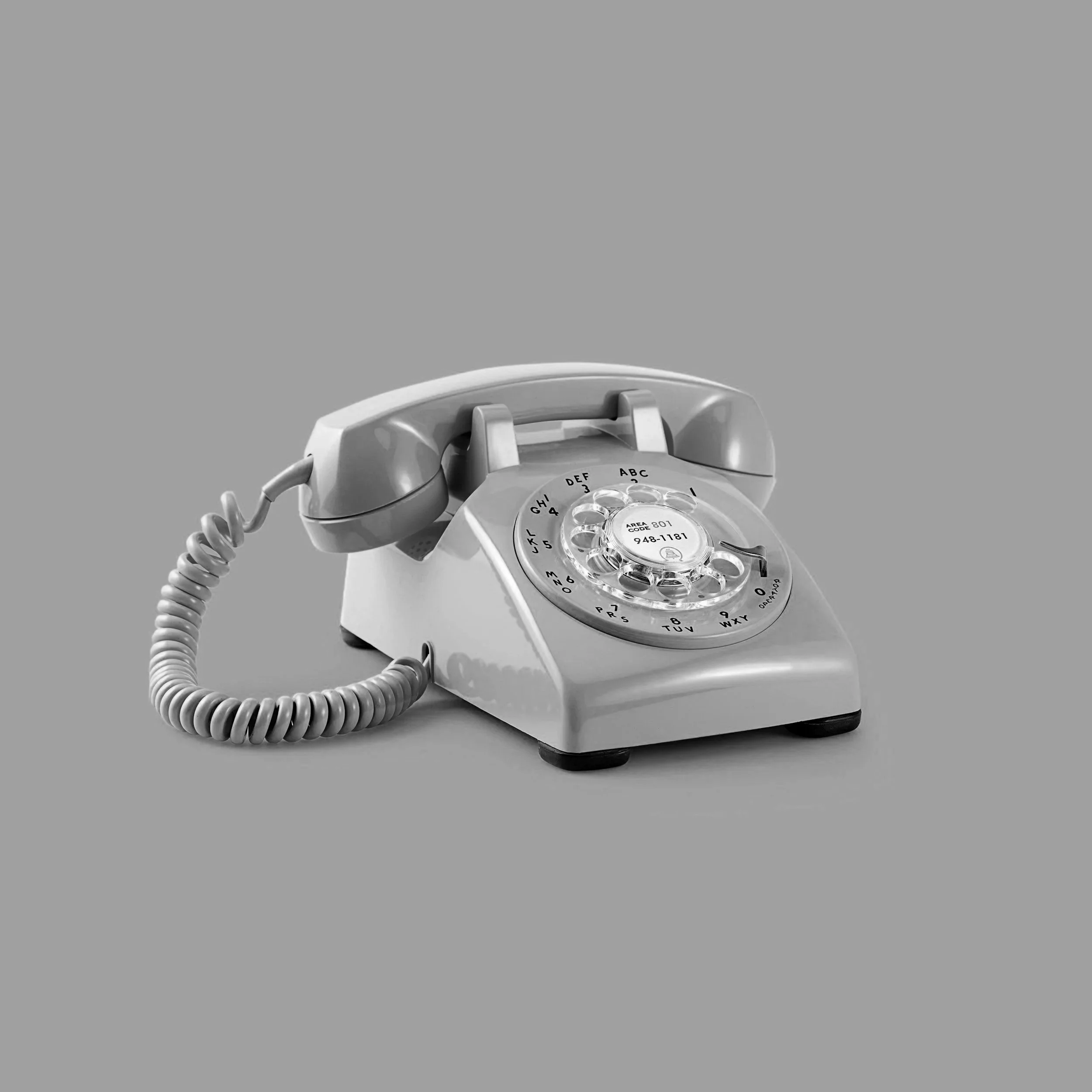 A rotary dial phone with a cord, gray in color, against a plain gray background.