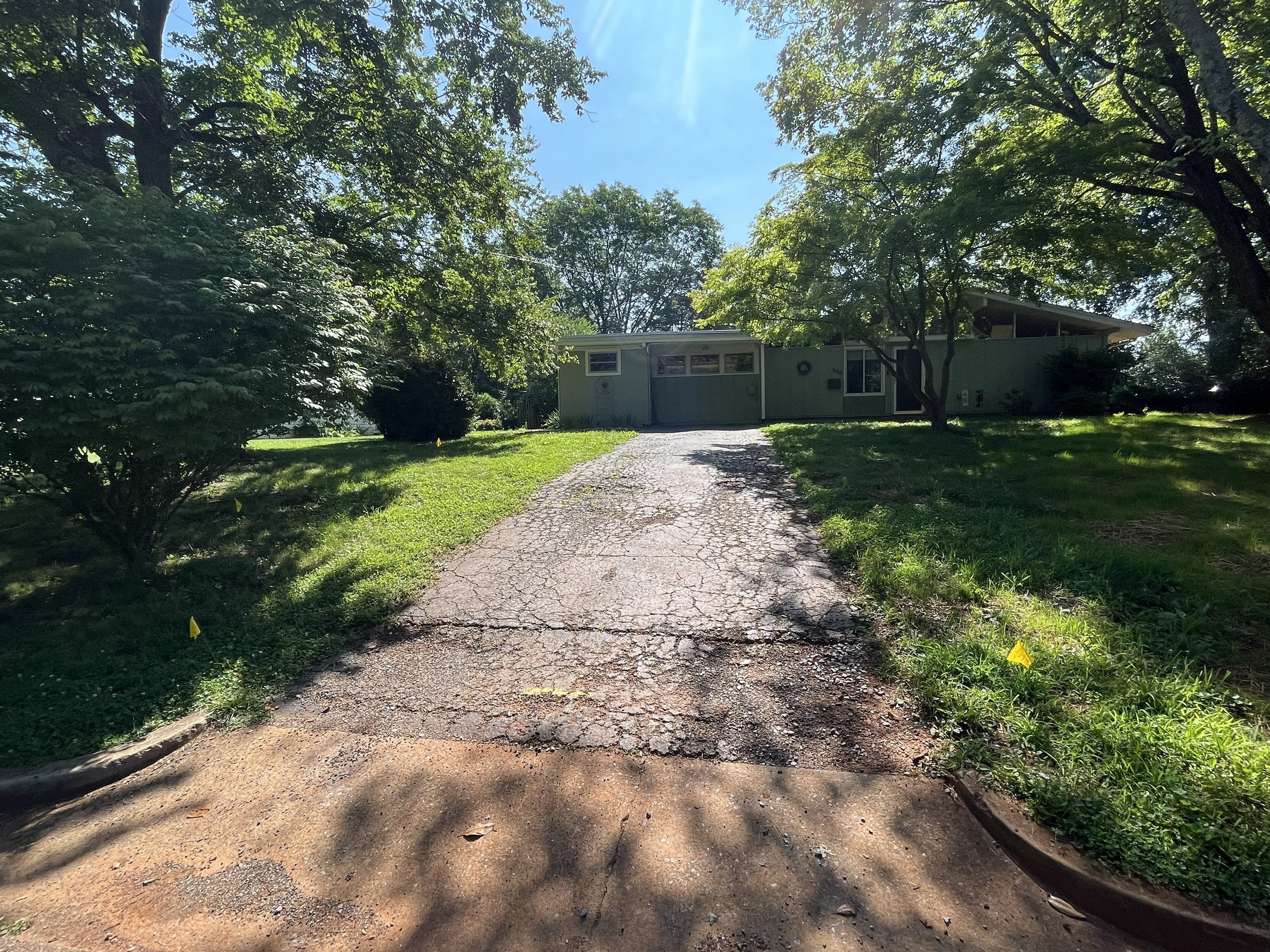Old and crumbling asphalt driveway on a busy residential street. The driveway is narrow and is bordered by overgown grass.