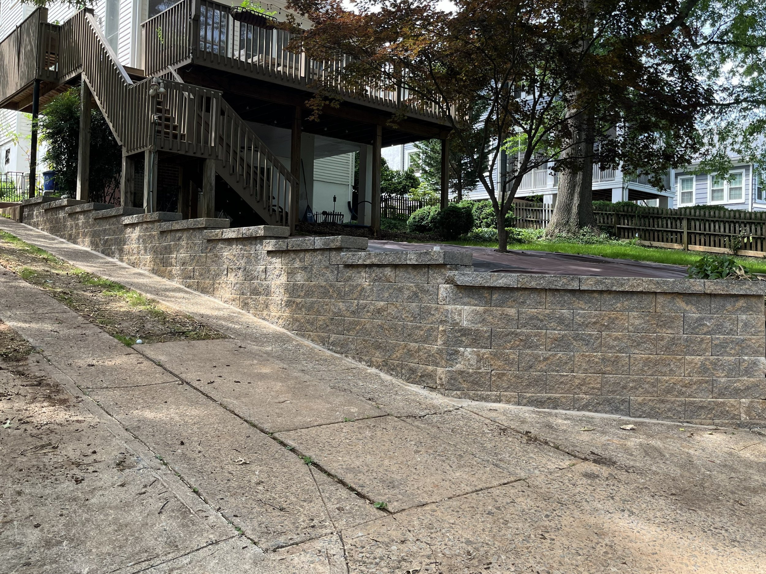 Newly constructed segmental block retaining wall next to a driveway that maximizes the yard and driveway space. The retaining wall is multi-tier and is a blend of dark browns and greys. All overgrown shrubs and bushes have been removed and replaced.