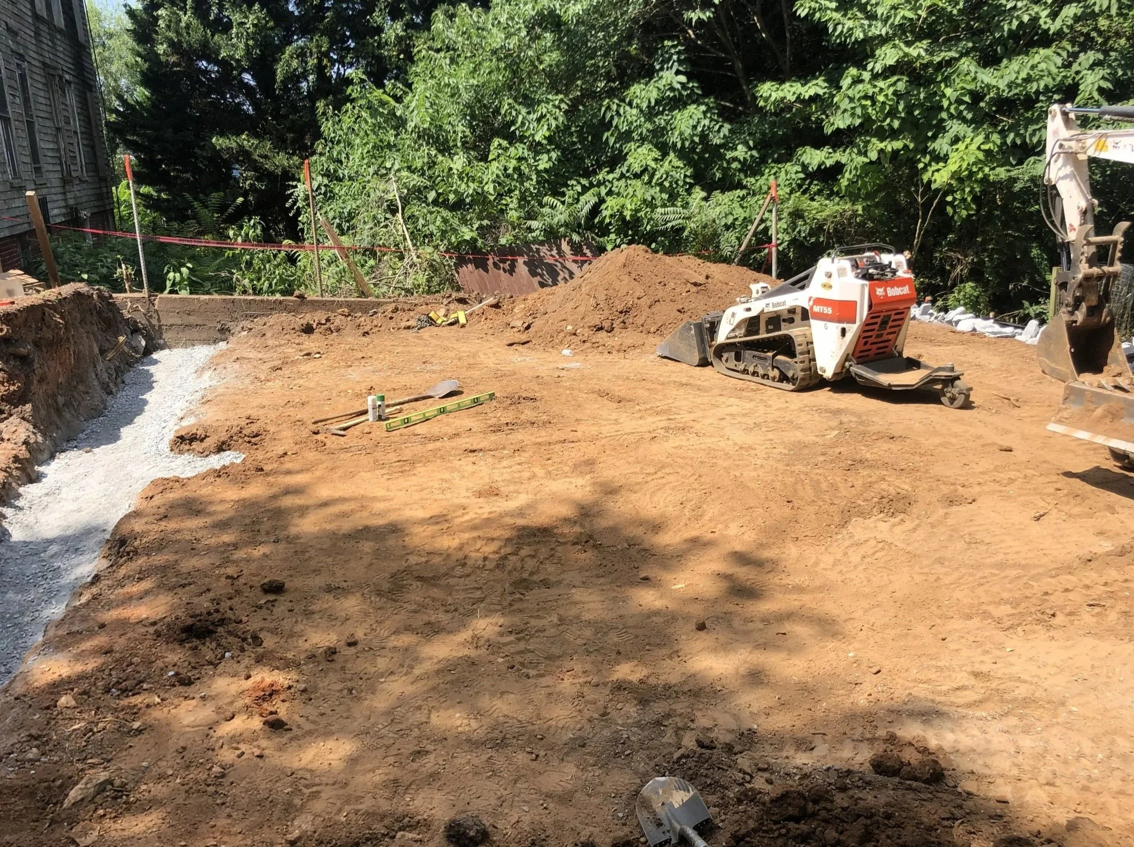 Construction site with dirt and excavation equipment, including a Bobcat compact loader, surrounding a partially dug area with a gravel pathway, and lush green trees in the background.
