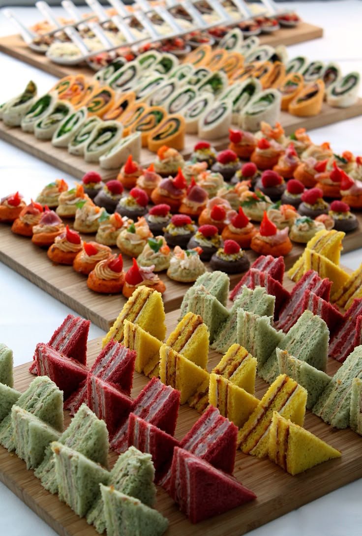 Assorted finger foods on wooden platters, including colorful mini sandwiches, canapés topped with vegetables and garnishes, and assorted small cake slices.