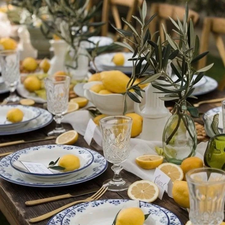 A table set for a meal with lemon-themed decor, including lemons on plates, in bowls, and as garnishes, with glassware, cutlery, and vases with greenery.