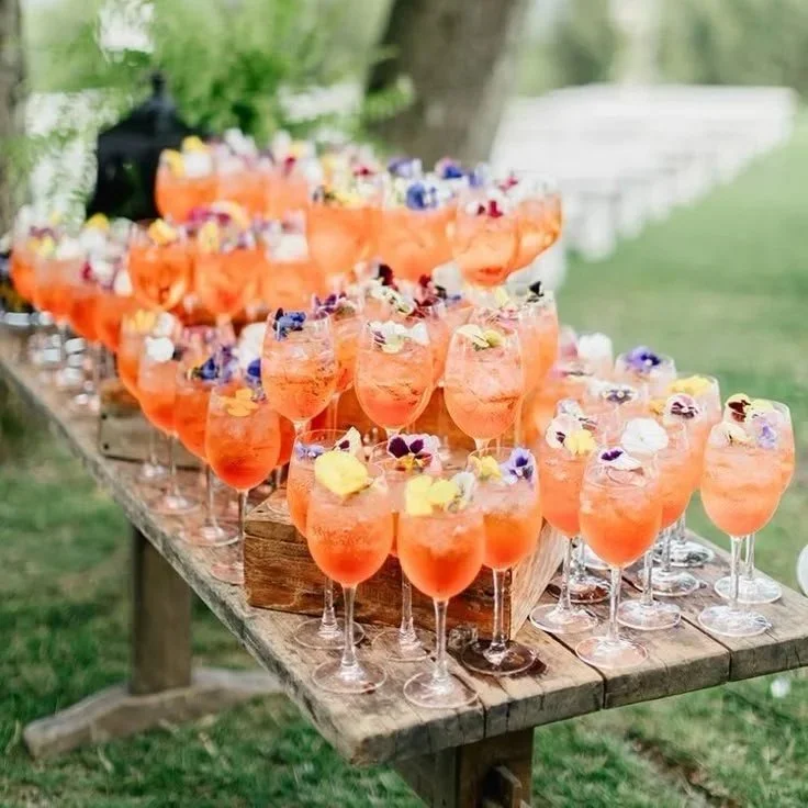 Several glasses of pink-orange cocktails garnished with edible flowers placed on a rustic wooden table outdoors.
