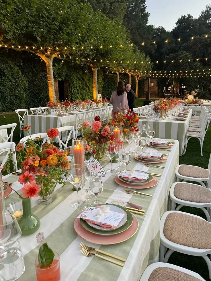 Outdoor evening wedding reception with long decorated tables, floral centerpieces, candles, and string lights overhead.