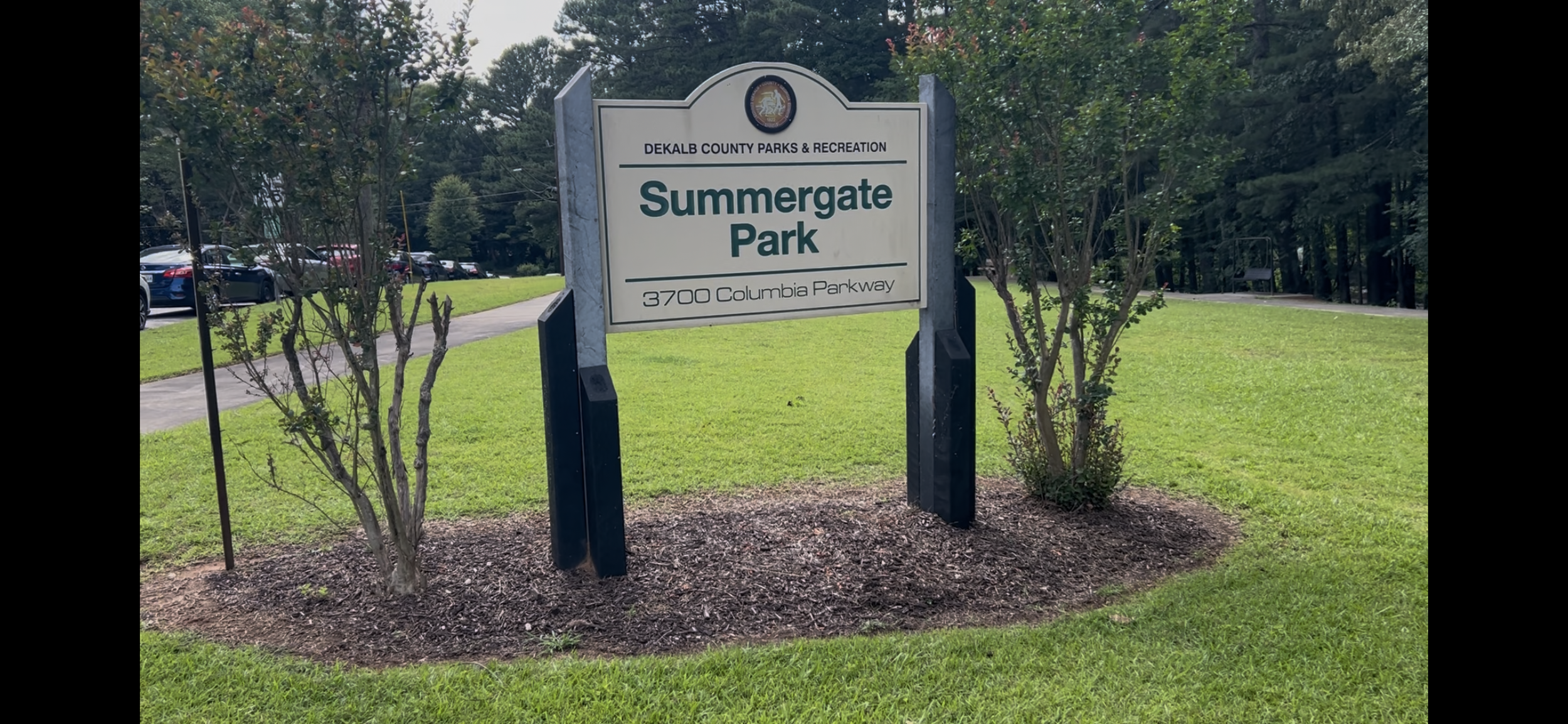 A sign for Summergate Park in DeKalb County, surrounded by green grass and small trees. The sign includes the park's address, 3700 Columbia Parkway, and the DeKalb County Parks & Recreation logo at the top.