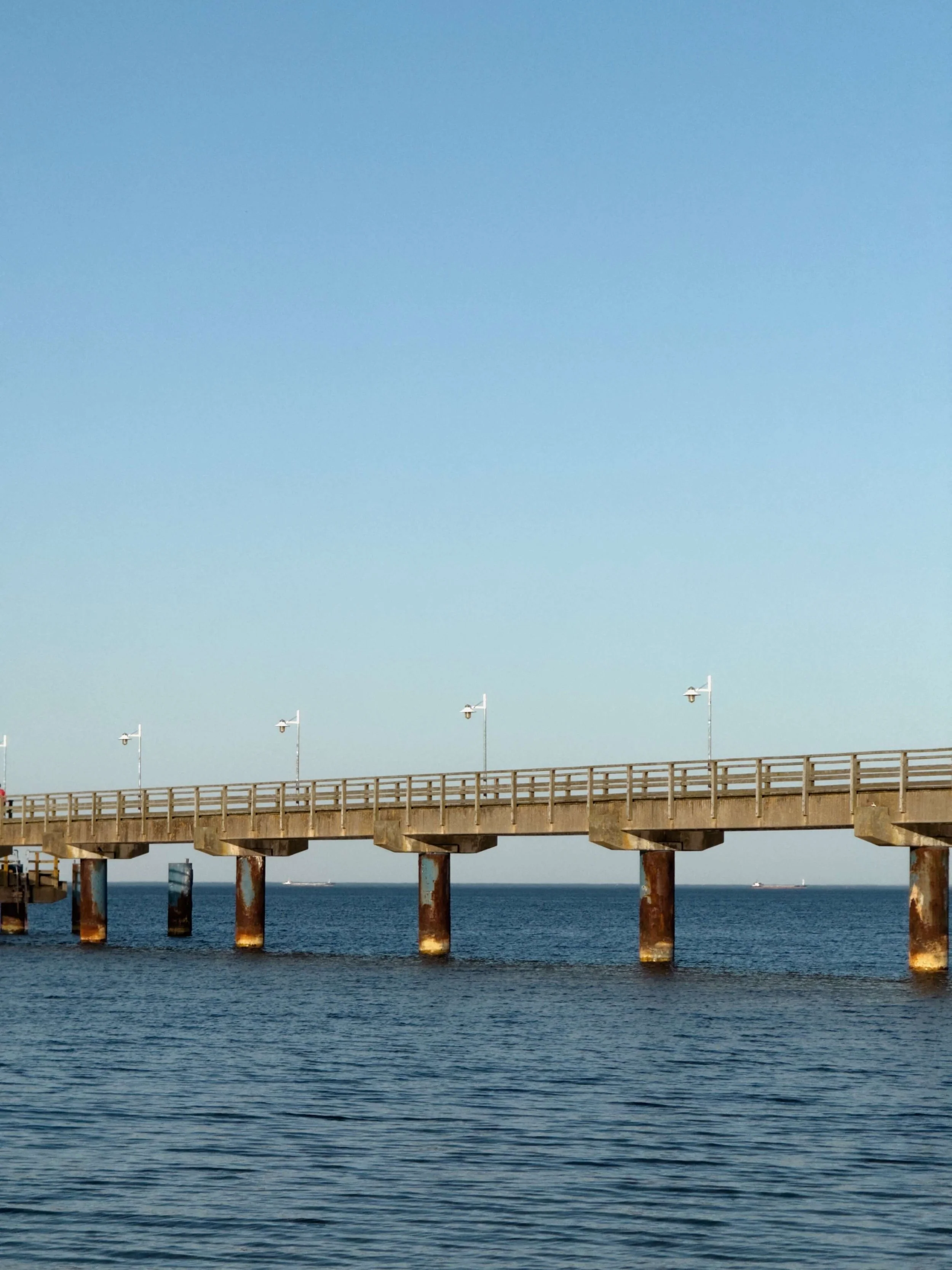 Ein Pier oder Steg erstreckt sich ins Meer, mit mehreren Laternen entlang der Seite, unter einem klaren blauen Himmel.