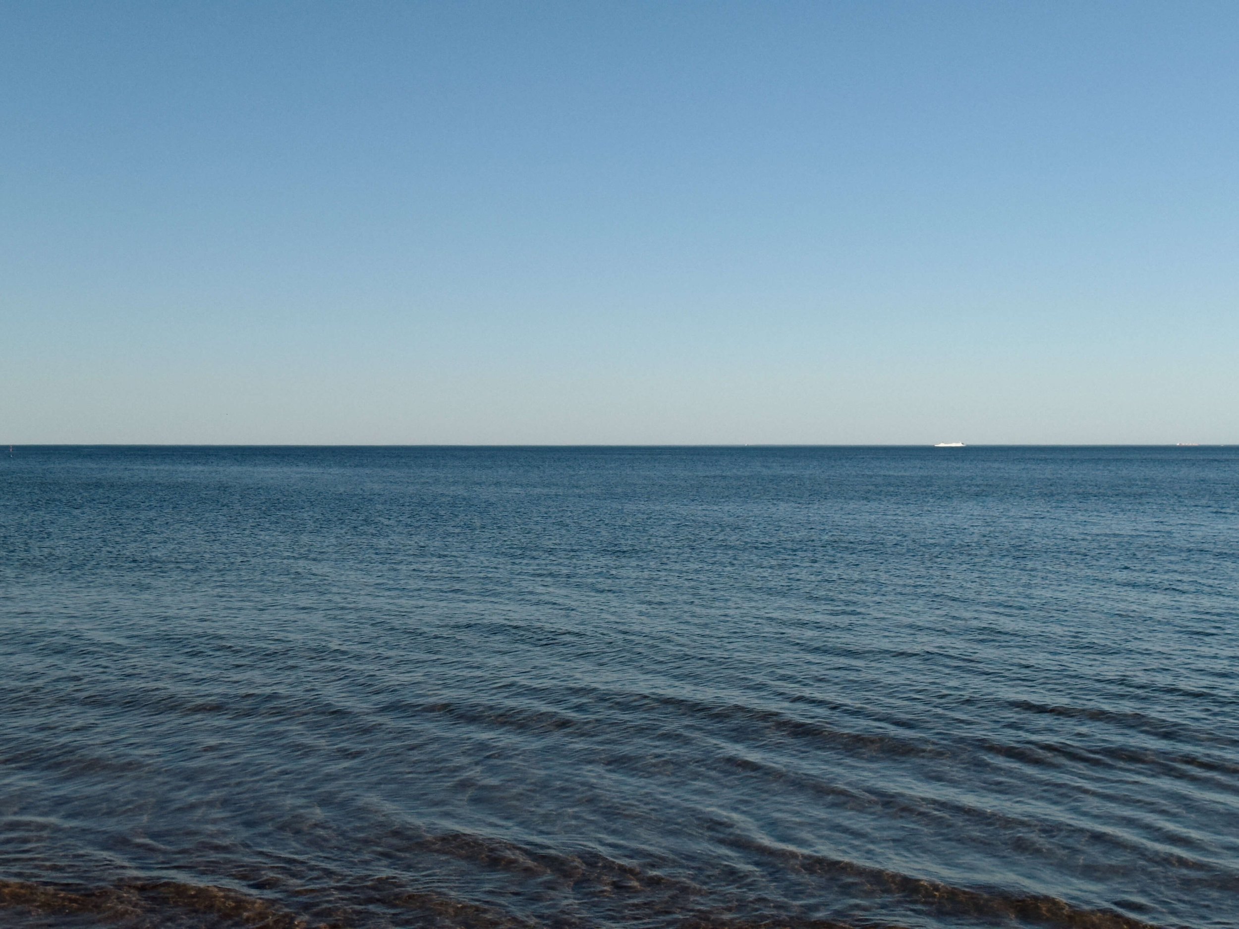 Blick auf das Meer unter einem klaren blauen Himmel, mit einem kleinen weißen Schiff im Horizont.