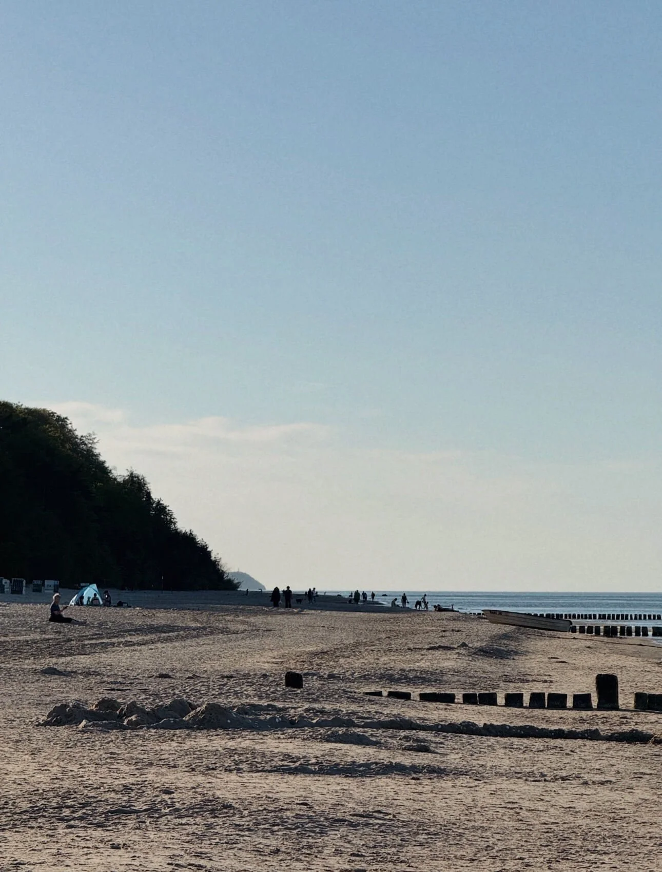 Ein Strand mit Menschen, die spazieren gehen oder sitzen, Bäumen auf der linken Seite, einem breiten Sandstrand und dem Meer im Hintergrund. Es ist wahrscheinlich später Nachmittag.