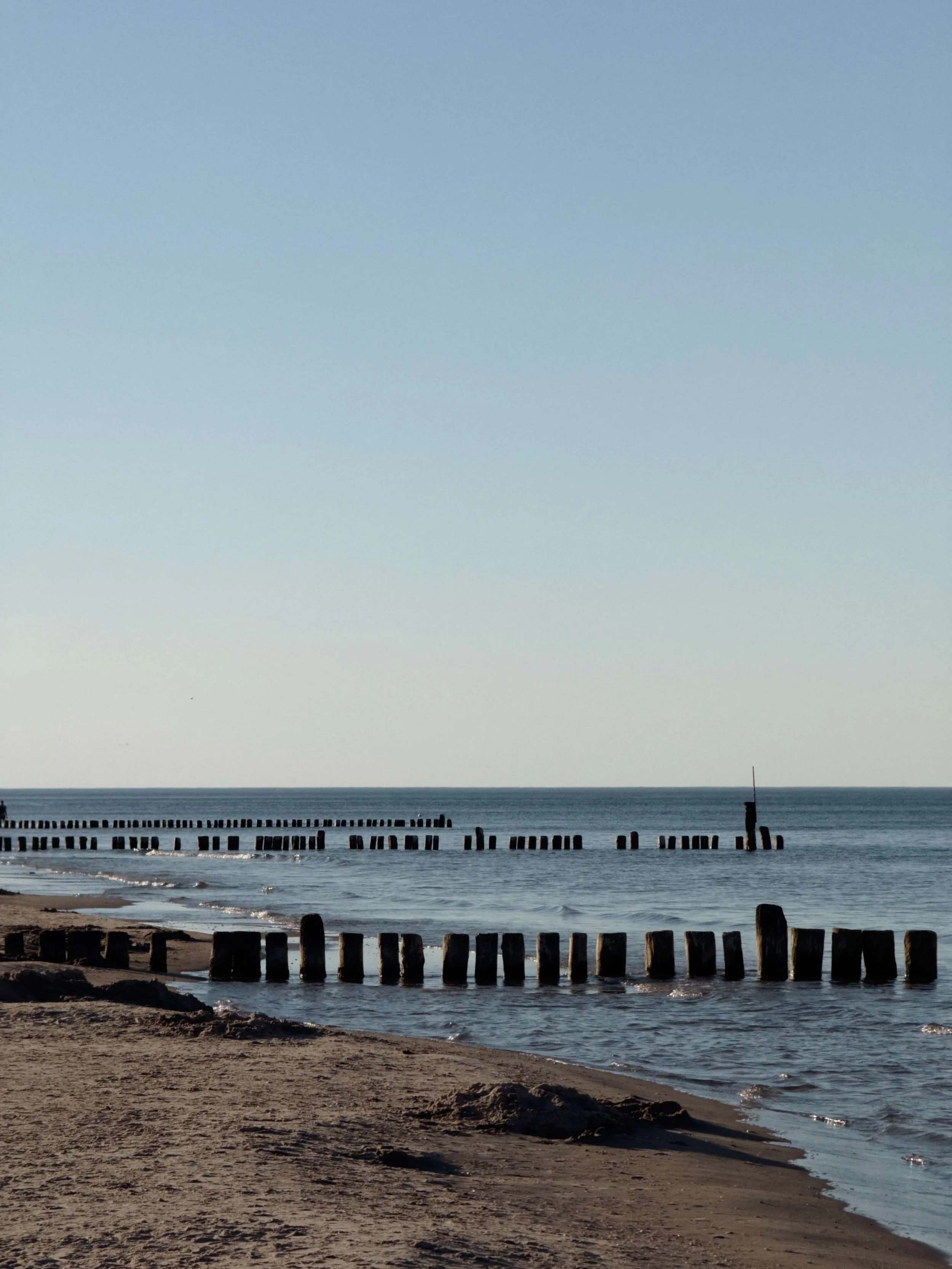 Ein Strand mit Holzpfählen im Wasser und im Hintergrund das offene Meer unter einem klaren Himmel.