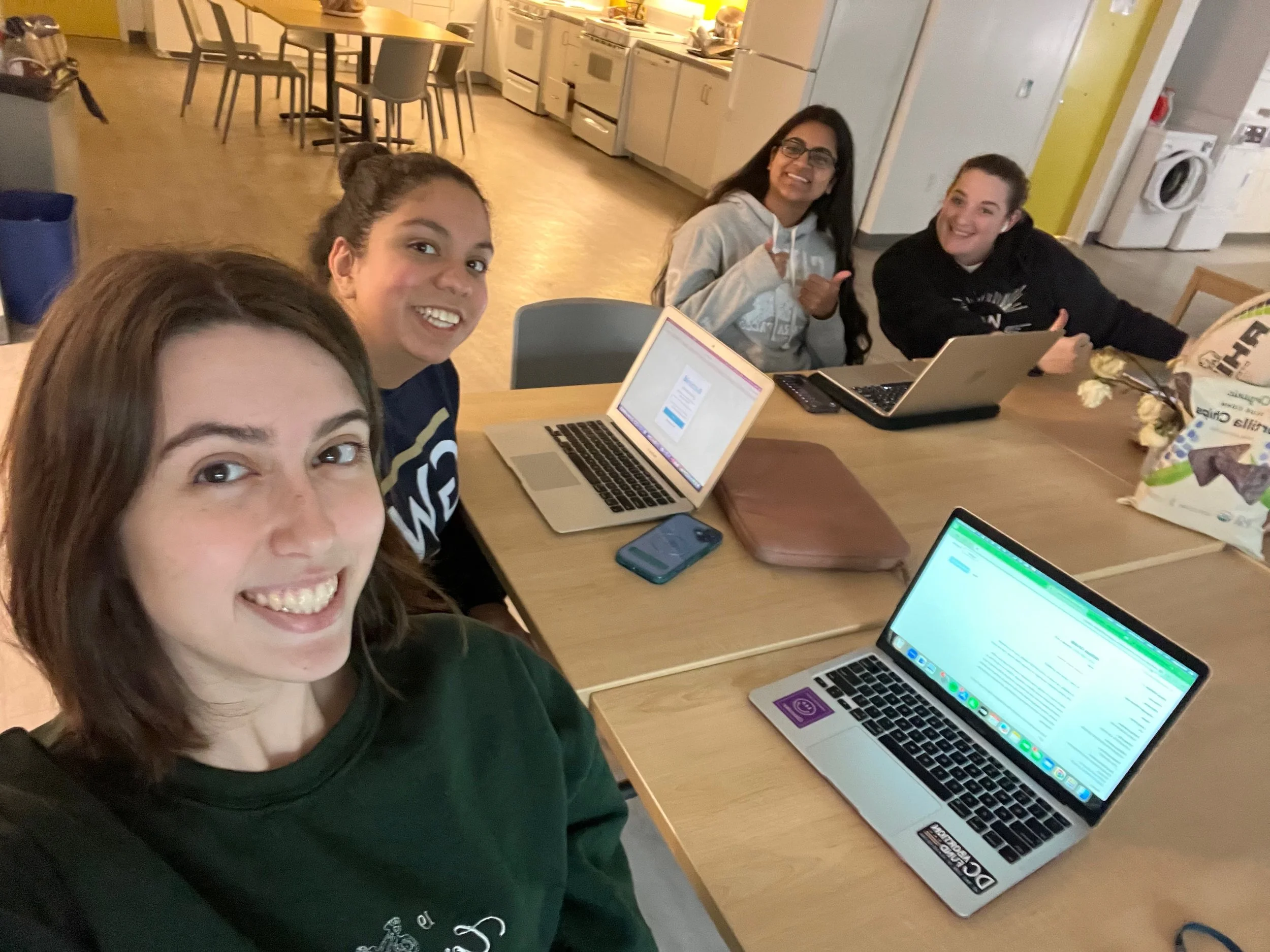 Four women with laptops sit around table smiling at camera