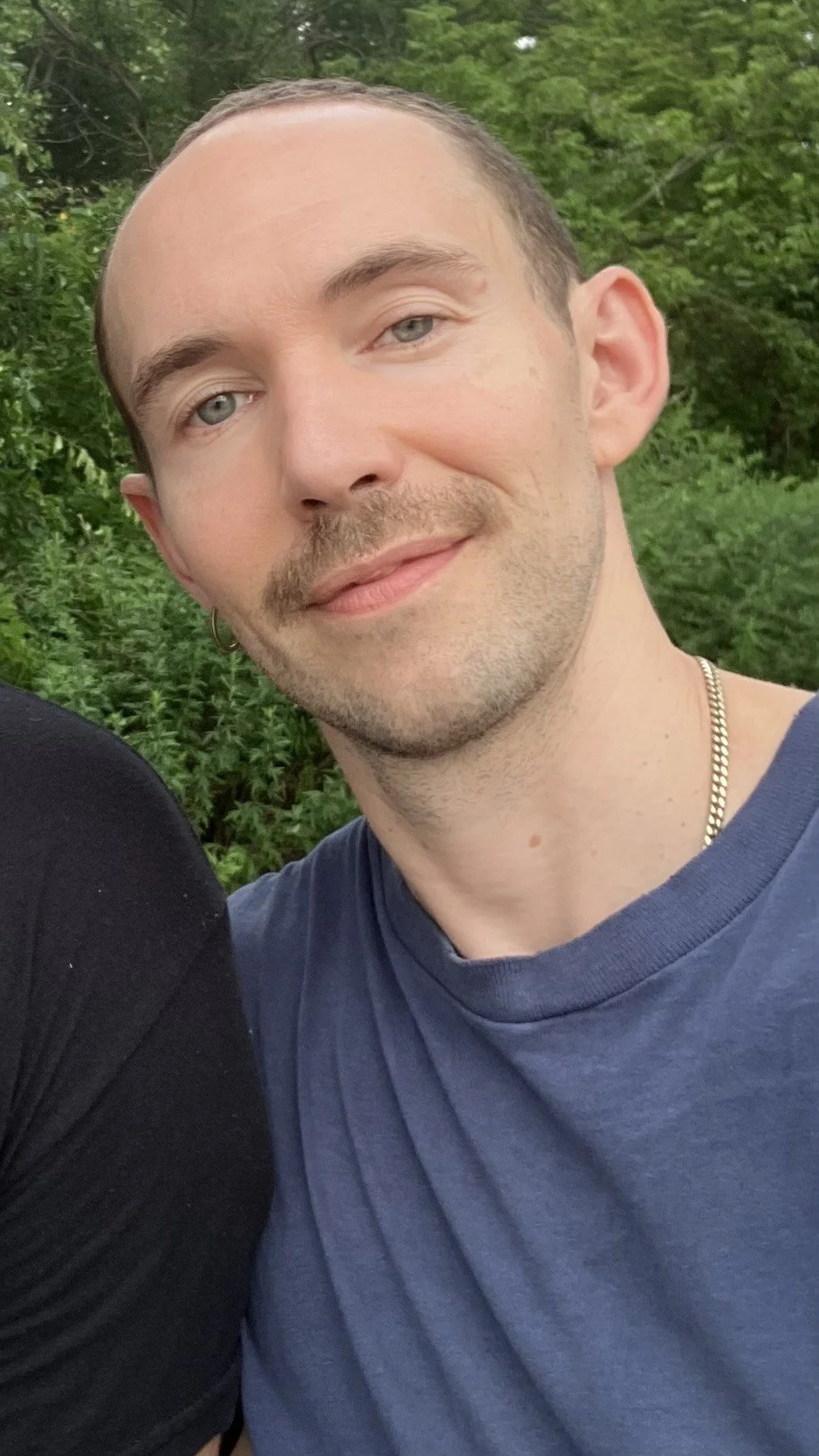 Close-up of a young man with light skin, blue eyes, short brown hair, a goatee, and a small hoop earring, smiling outdoors with green foliage in the background.