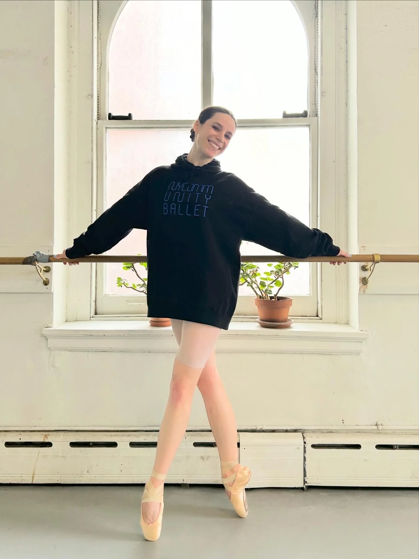 A ballerina standing in front of a large window with white walls, wearing a black sweatshirt with 'HUMM UNITI BALLET' written on it, pink ballet tights, and pointe shoes, smiling and posing with arms outstretched holding a wooden ballet barre.