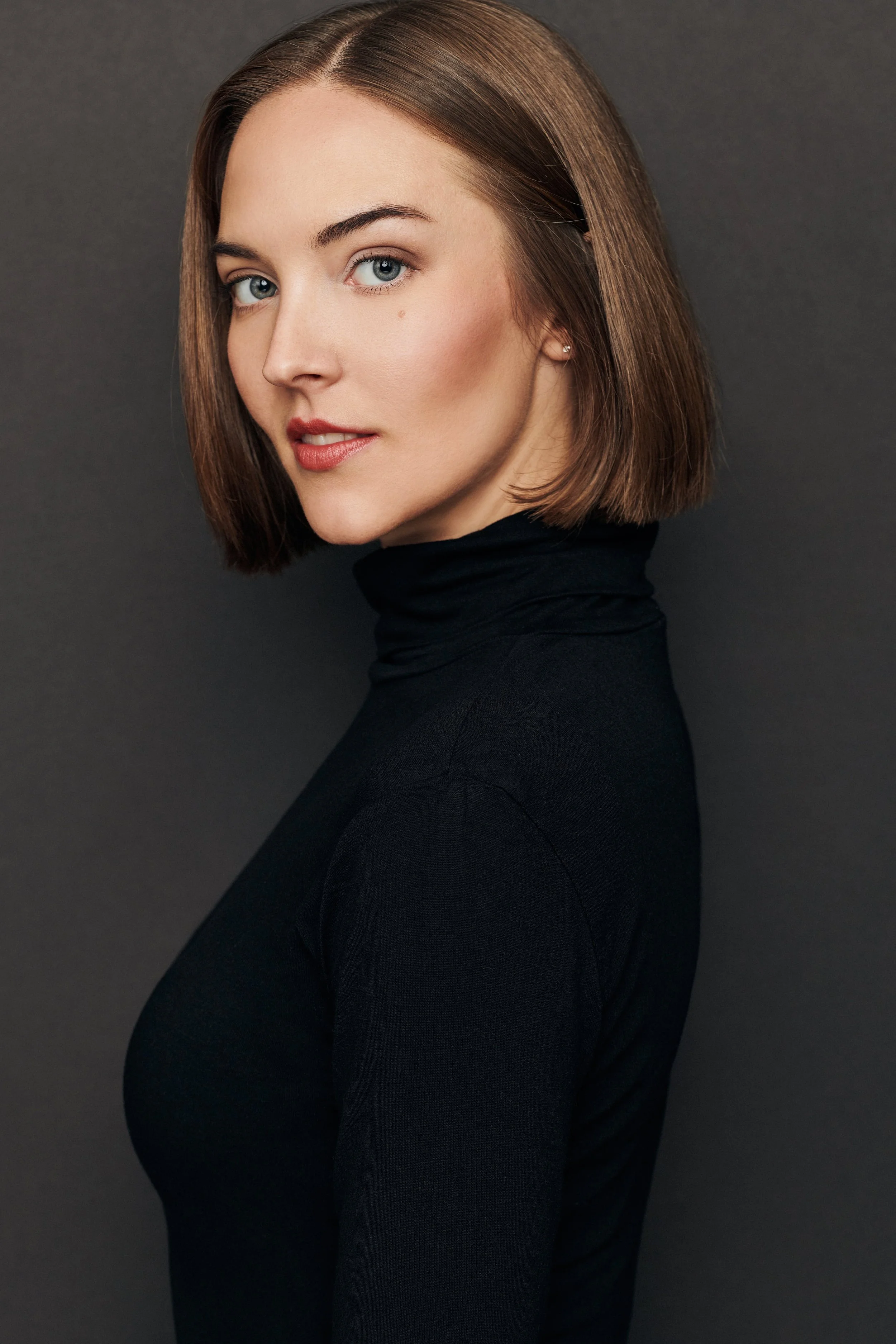 A portrait of a young woman with shoulder-length brown hair, blue eyes, and fair skin, wearing a black turtleneck and small earrings, against a dark background.
