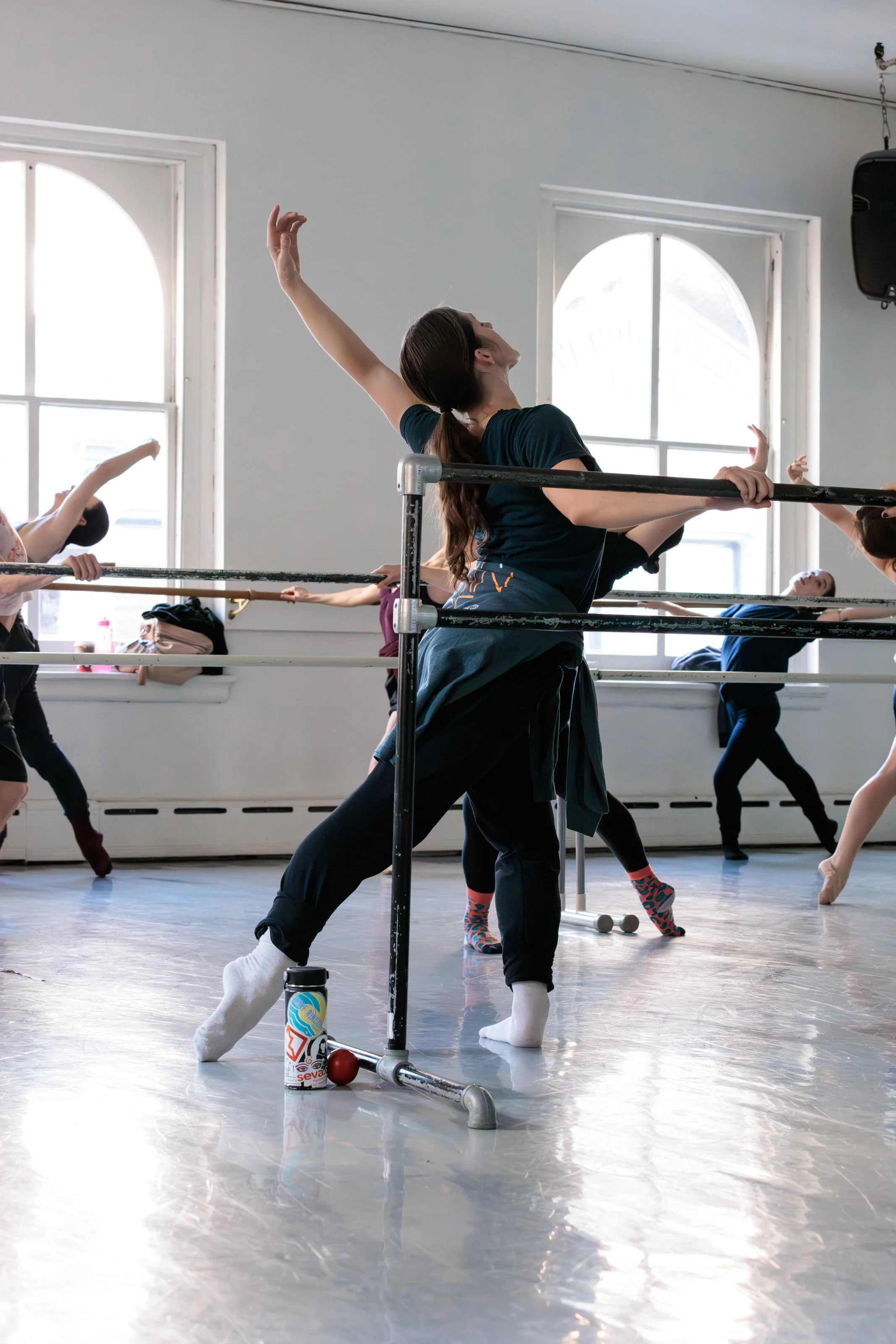 Dance class with multiple dancers practicing ballet at barre, with a woman in the foreground stretching. Bright room with large windows and wooden floor.