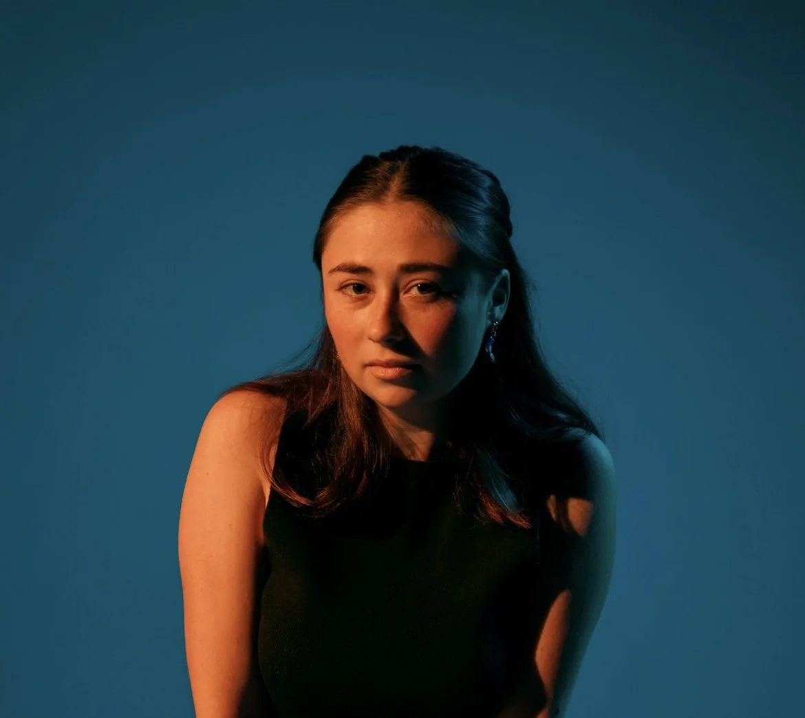 A young woman with long brown hair posed against a gradient blue background, wearing a black sleeveless top.