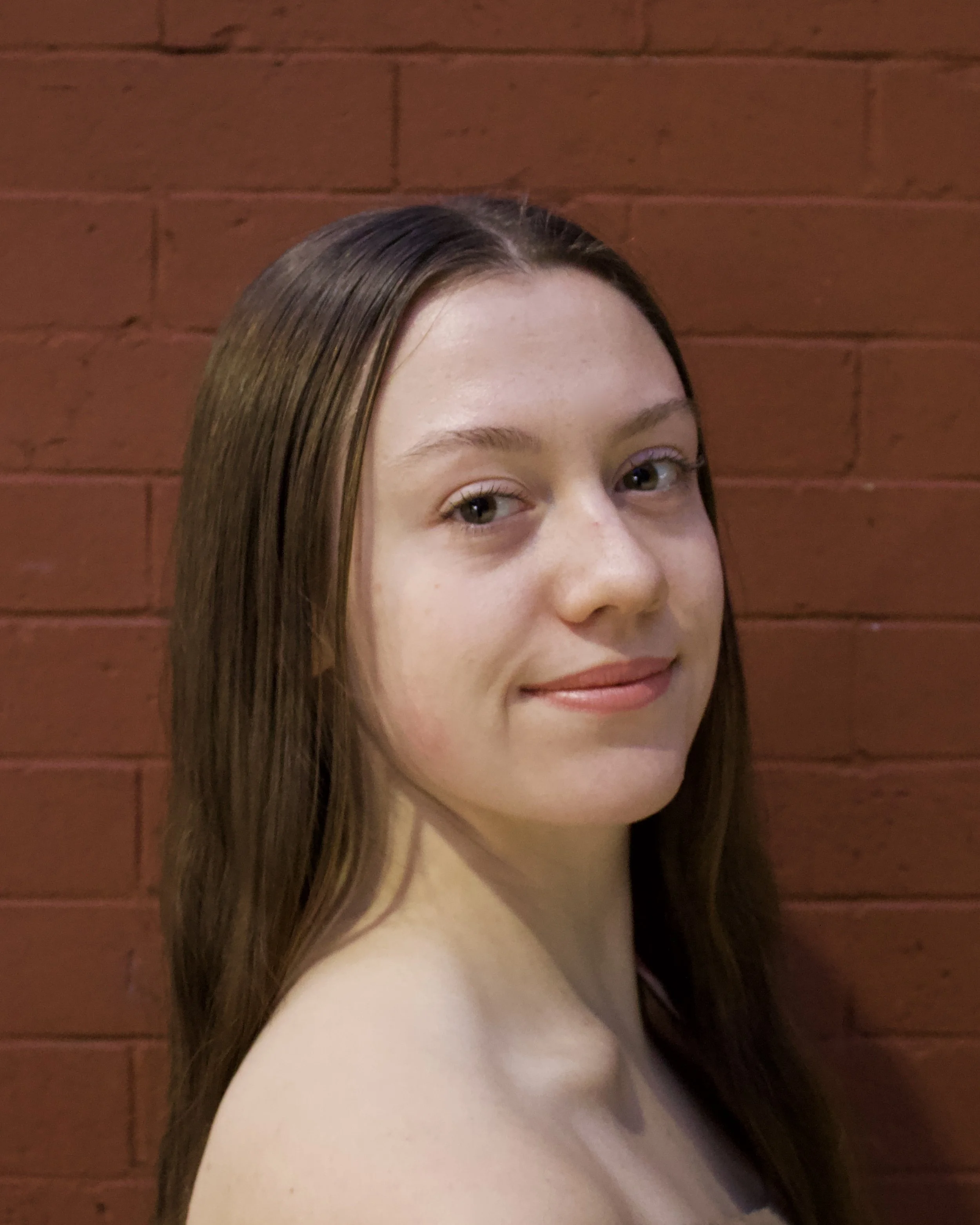 A young woman with long brown hair and fair skin standing against a brick wall.