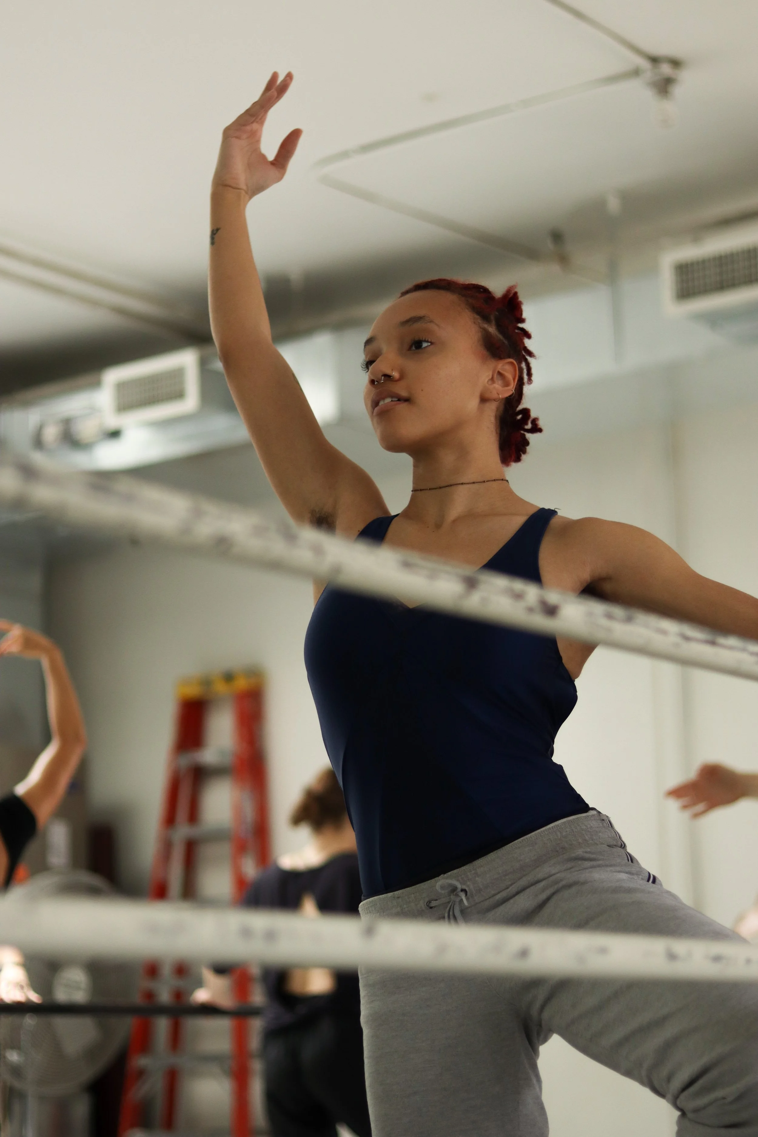 A woman with reddish hair in a dance studio performs a dance move with her right arm raised above her head. She wears a navy blue tank top and grey sweatpants, and is focused on her movements. Other dancers are visible in the background.