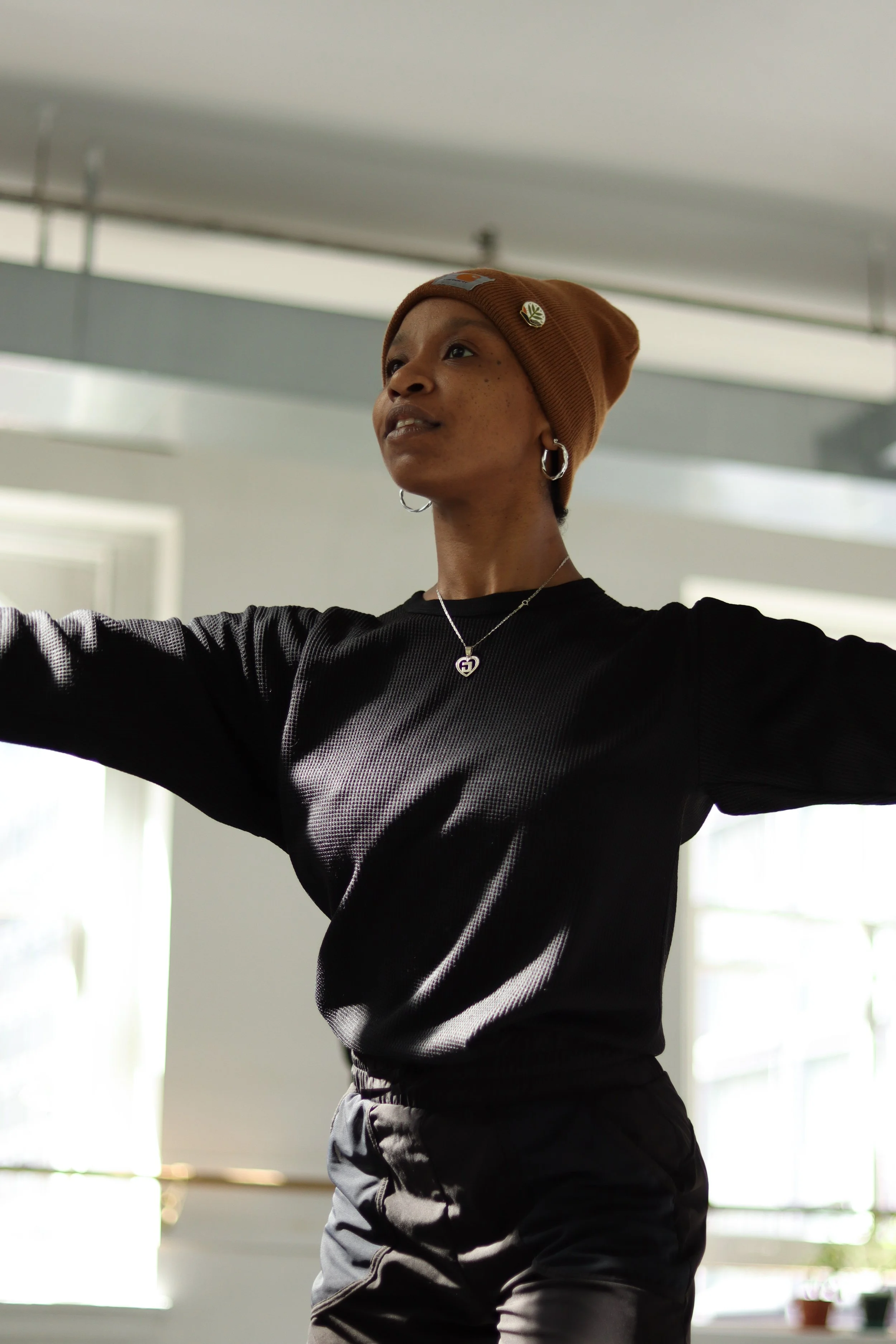 A woman wearing a brown beanie hat with pins, hoop earrings, a necklace, and a black athletic top, standing indoors with arms raised.