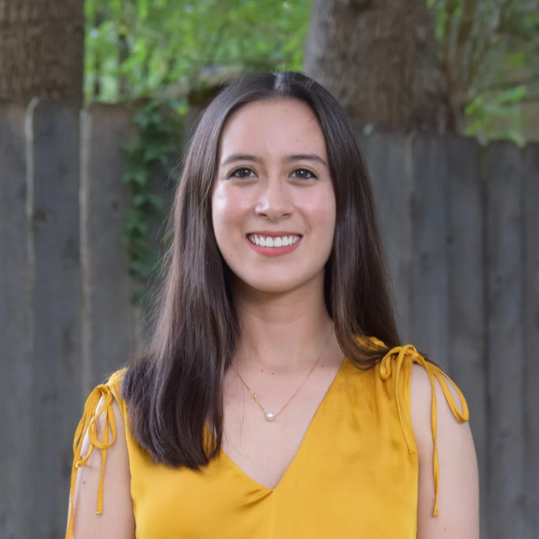 A smiling young woman with long dark hair, wearing a yellow sleeveless top with tie straps, standing outdoors in front of a wooden fence and leafy trees.