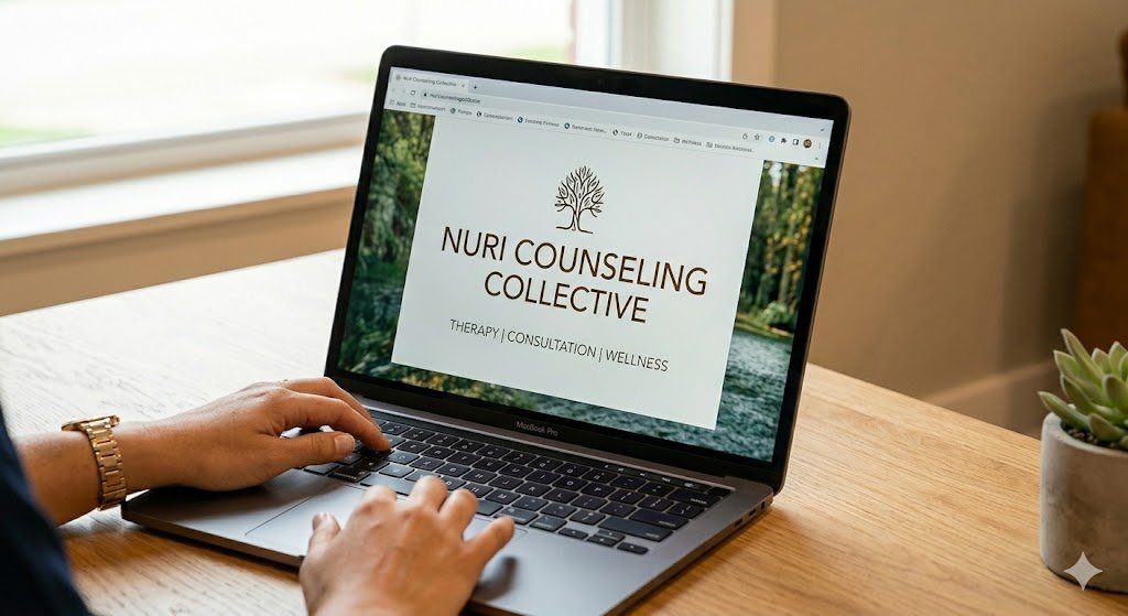 Laptop displaying Nuri Counseling Collective website with a logo of a tree, on a wooden table with a person using it, near a window with sunlight, and a potted succulent plant nearby.
