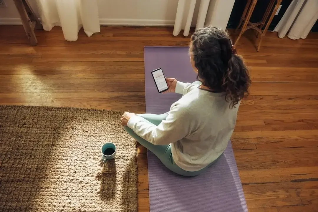 A woman with curly hair sitting on a purple yoga mat on a wooden floor, holding a smartphone in her right hand and a coffee mug nearby, with curtains and a small table in the background.