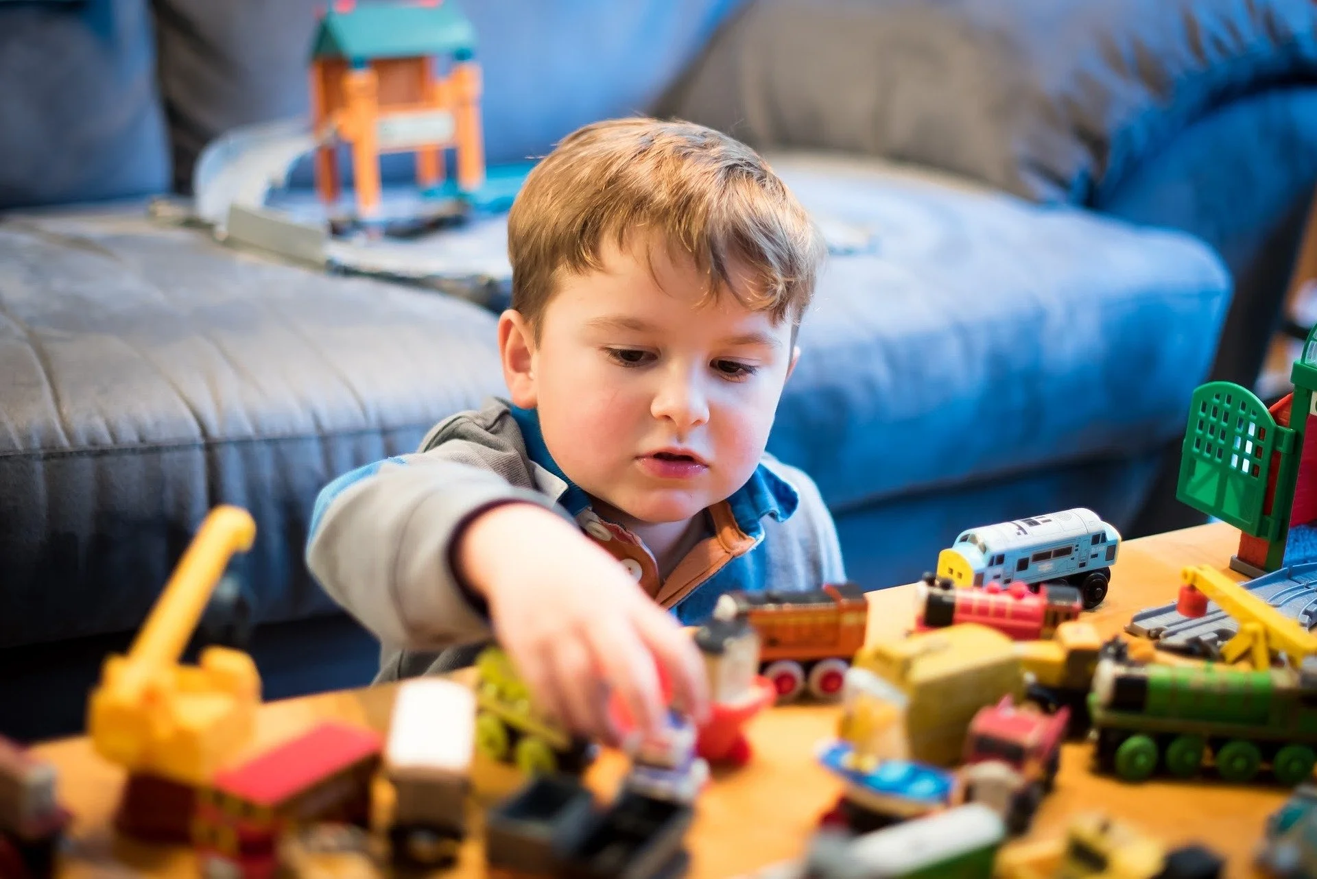 A young boy with brown hair playing with toy trains on a table in a living room. There are toy buildings and accessories around him, with a gray sofa in the background.