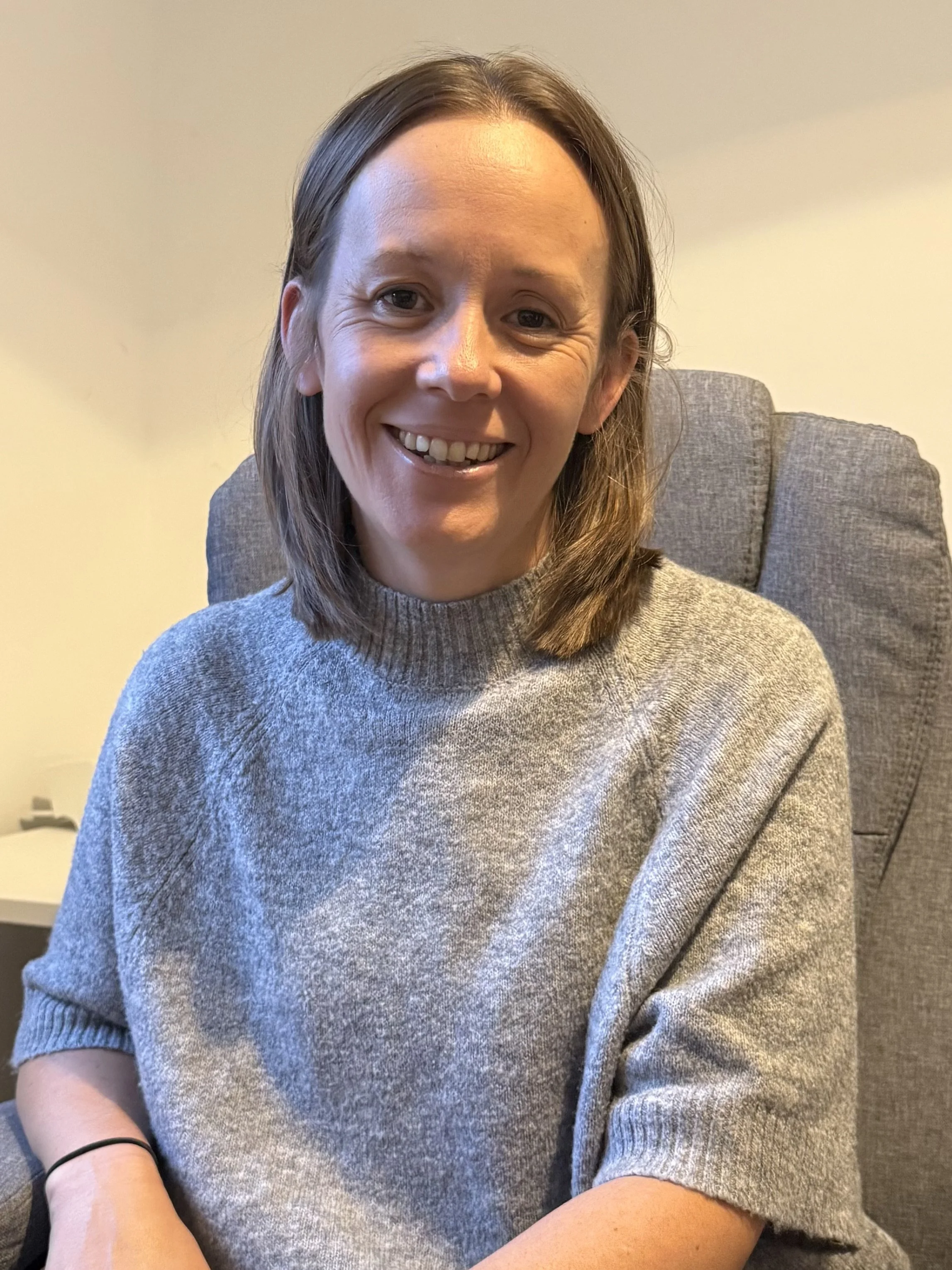 A woman with shoulder-length brown hair wearing a gray sweater, sitting in a beige-colored room on a gray chair, smiling at the camera.