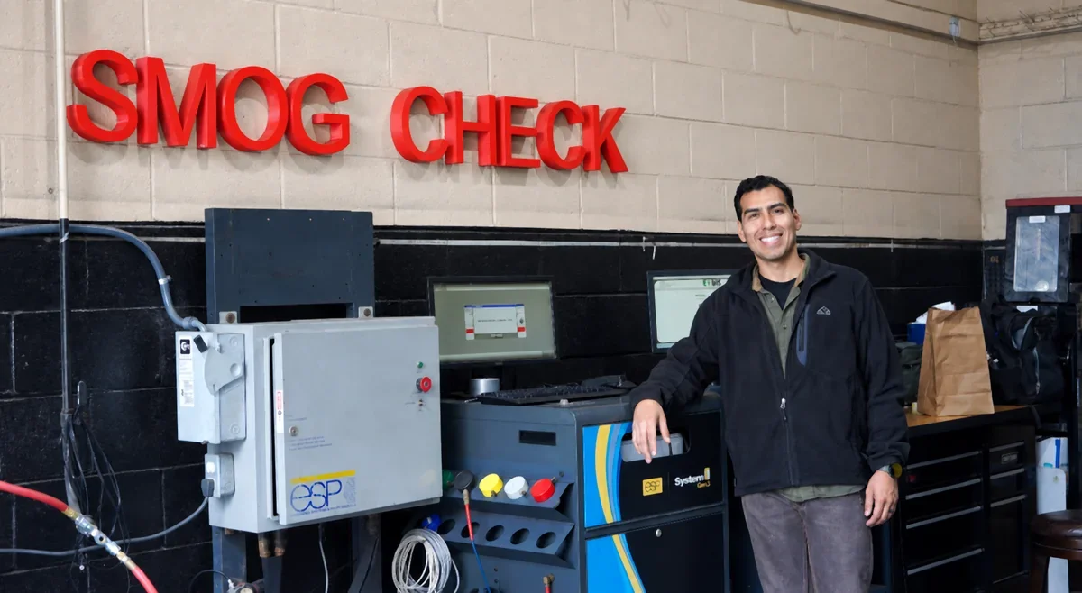 OC Smog Lab technician standing beside the indoor red Smog Check sign and emissions-testing equipment