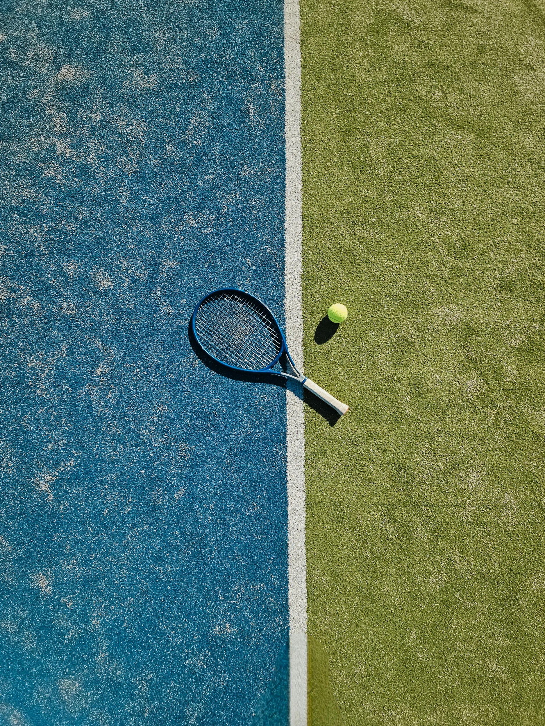 A tennis court with two distinct surfaces, one blue and one green, separated by a white line. A tennis racket and a tennis ball rest on the court.