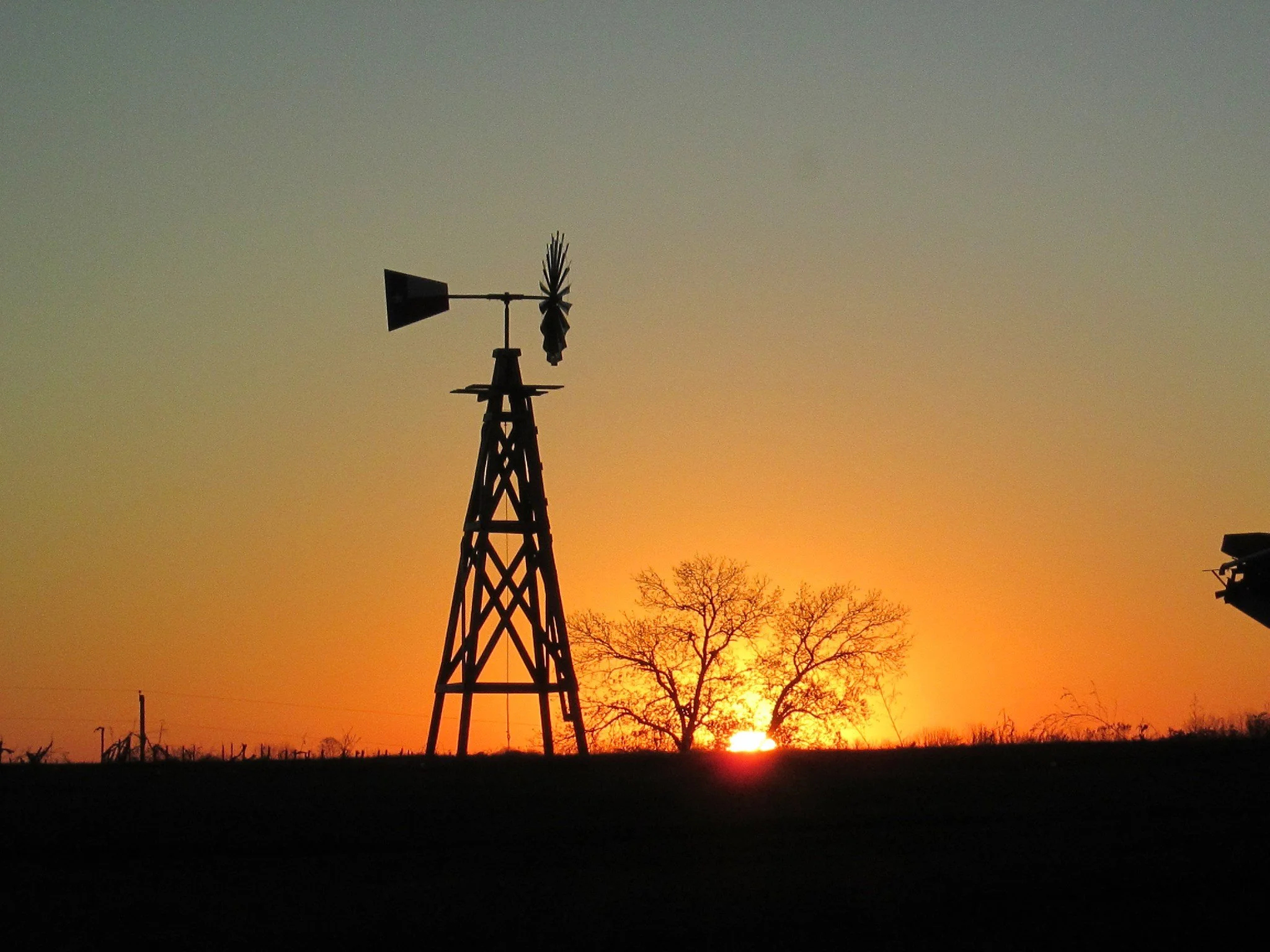 Silhouette of a windmill structure and trees against a sunset sky with shades of orange and yellow.
