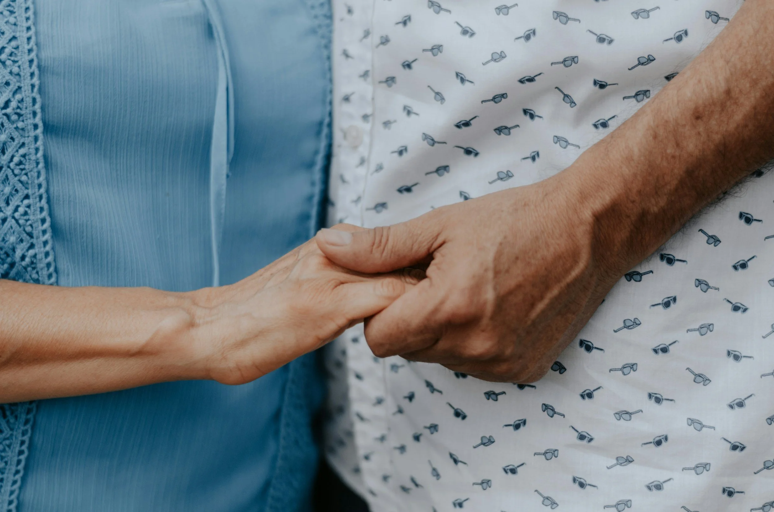 Close-up of two hands holding each other, one is elderly and the other appears middle-aged, set against hospital or medical gown fabric with a pattern of small glasses.