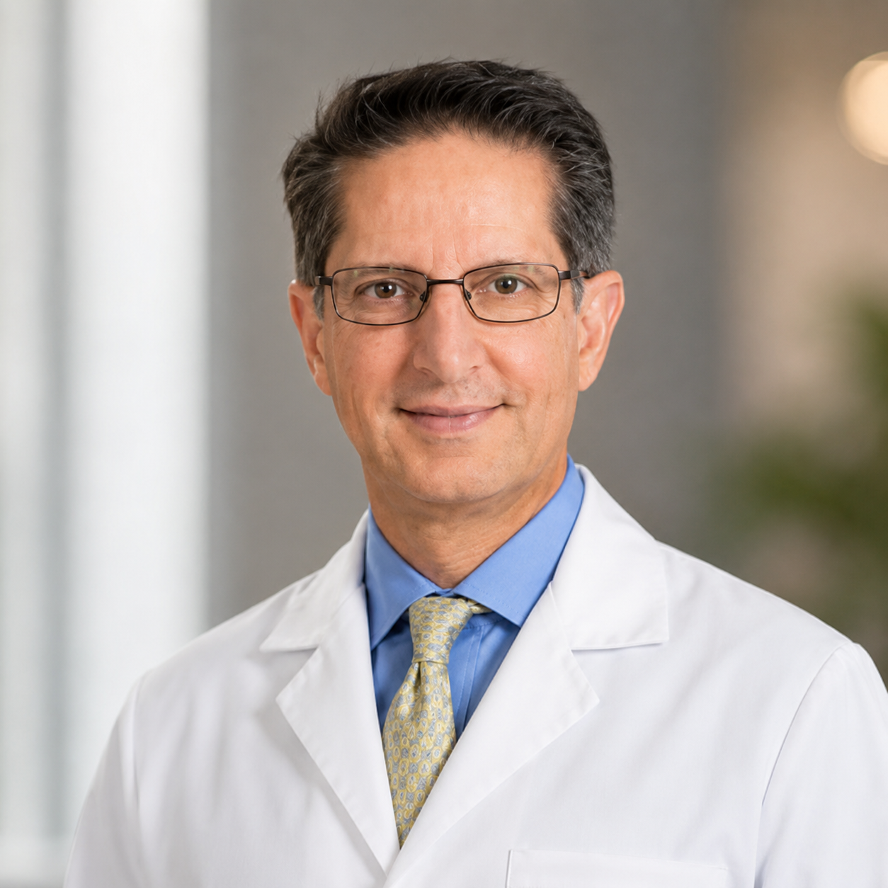 Male doctor wearing glasses, white coat, blue shirt, and yellow tie, smiling indoor.