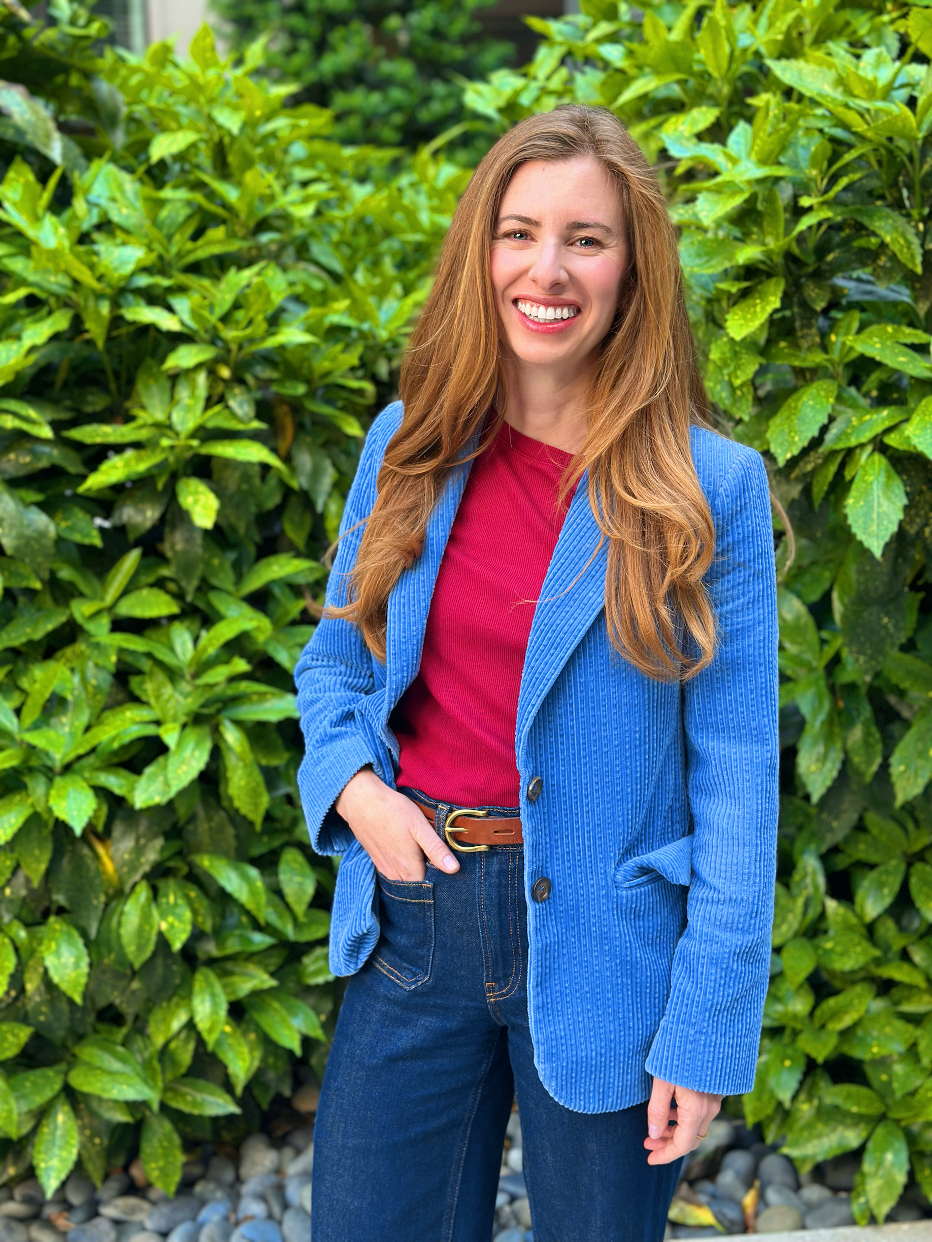 A woman with long red hair wearing a red top, a blue blazer, and blue jeans standing outdoors in front of green foliage.
