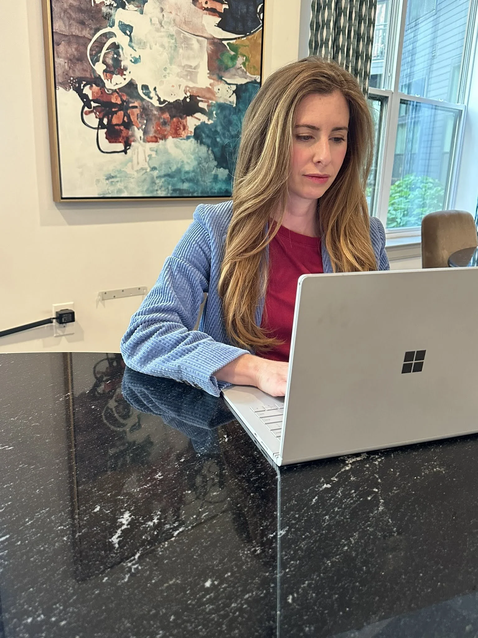 A woman with long, wavy, light brown hair working on a silver laptop at a black marble table in a bright room with large windows, colorful abstract artwork on the wall, and patterned curtains.