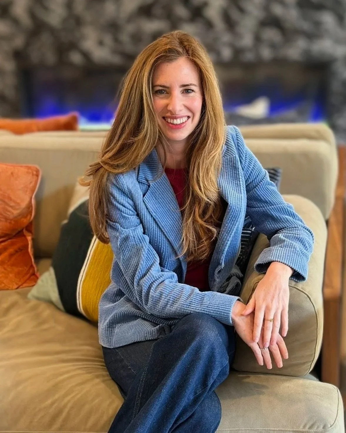 A woman with long, wavy red hair smiling while sitting on a couch in a cozy living room with a stone fireplace in the background.