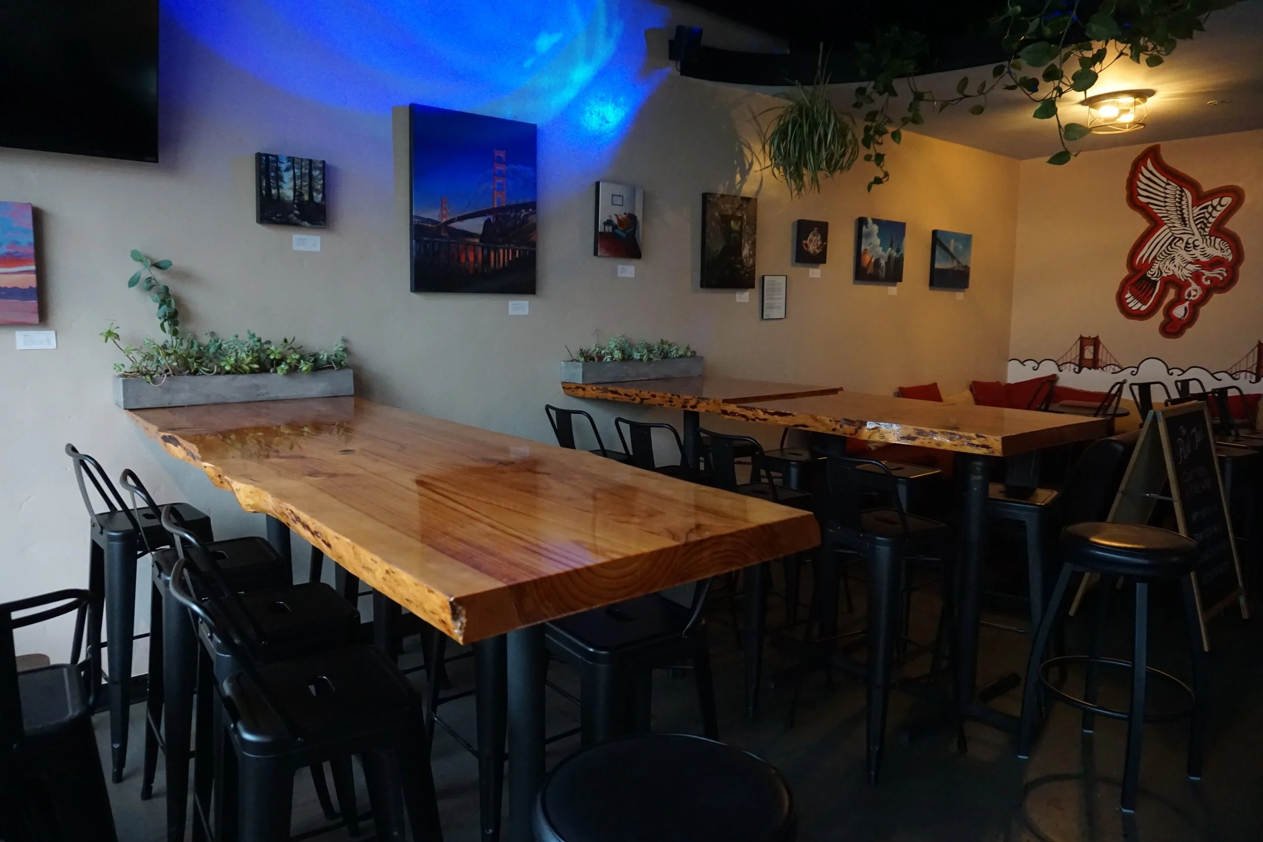 Interior of a restaurant or cafe with wooden tables, black chairs, and wall art including paintings and a large dragon mural. There are plants hanging from the ceiling and small potted plants on the tables. Dim lighting creates a cozy atmosphere.