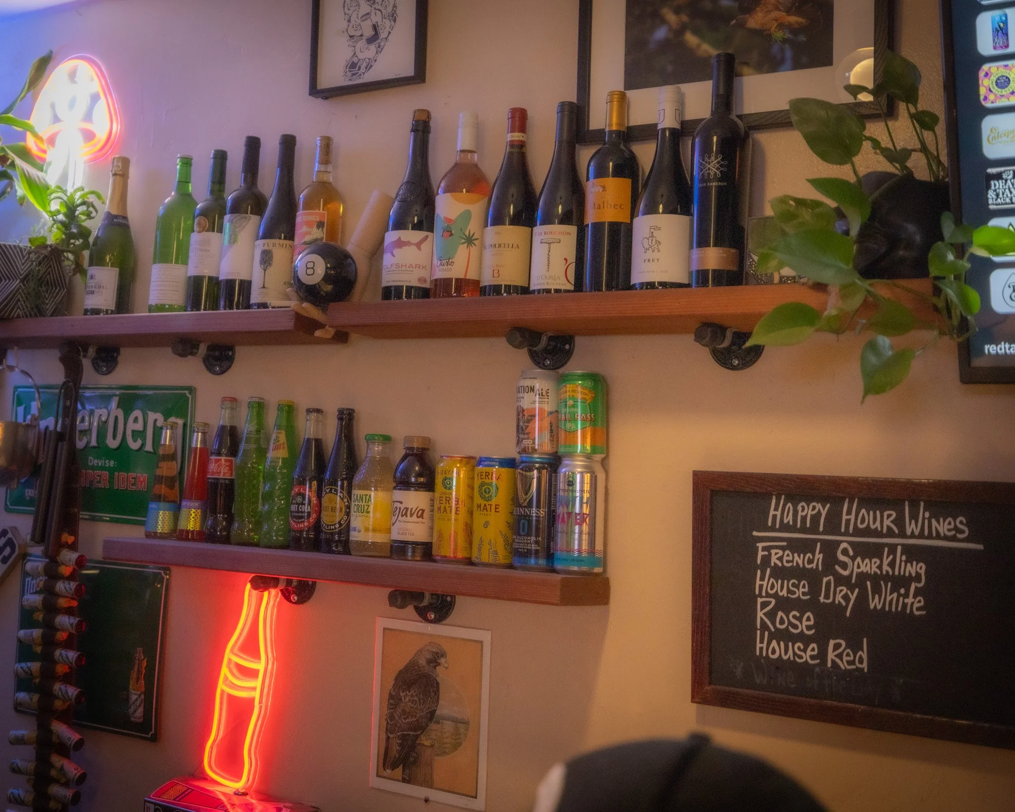 A wooden shelf on a beige wall holding various bottles of wine, with framed artwork, a neon sign, and a plant on either side. A chalkboard menu lists wines available for happy hour.