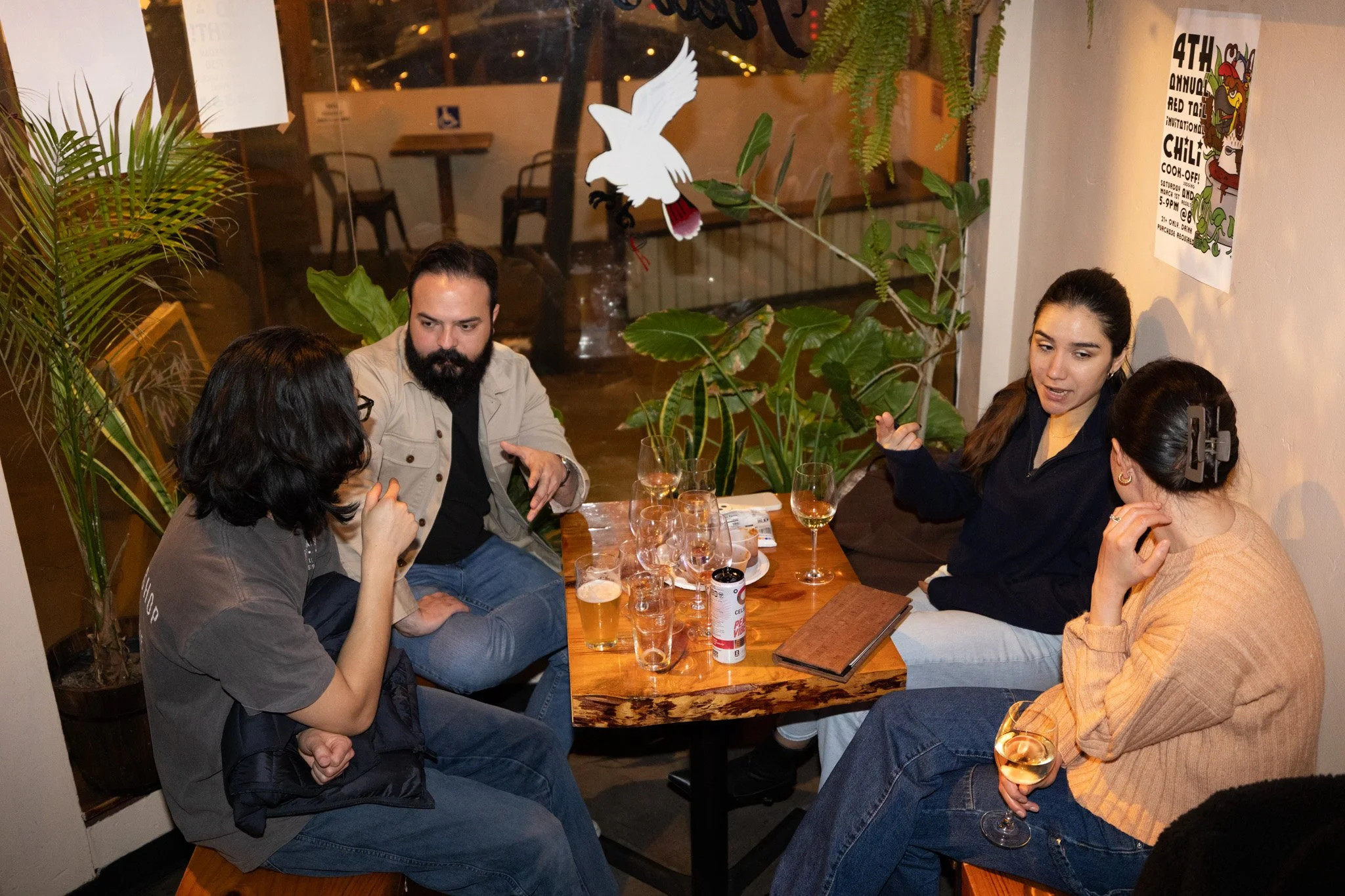 A group of five people sitting around a wooden table in a cozy restaurant, engaged in conversation. There are drinks on the table, including glasses of wine and beer. The setting is decorated with plants and a paper bird hanging from the ceiling. A p