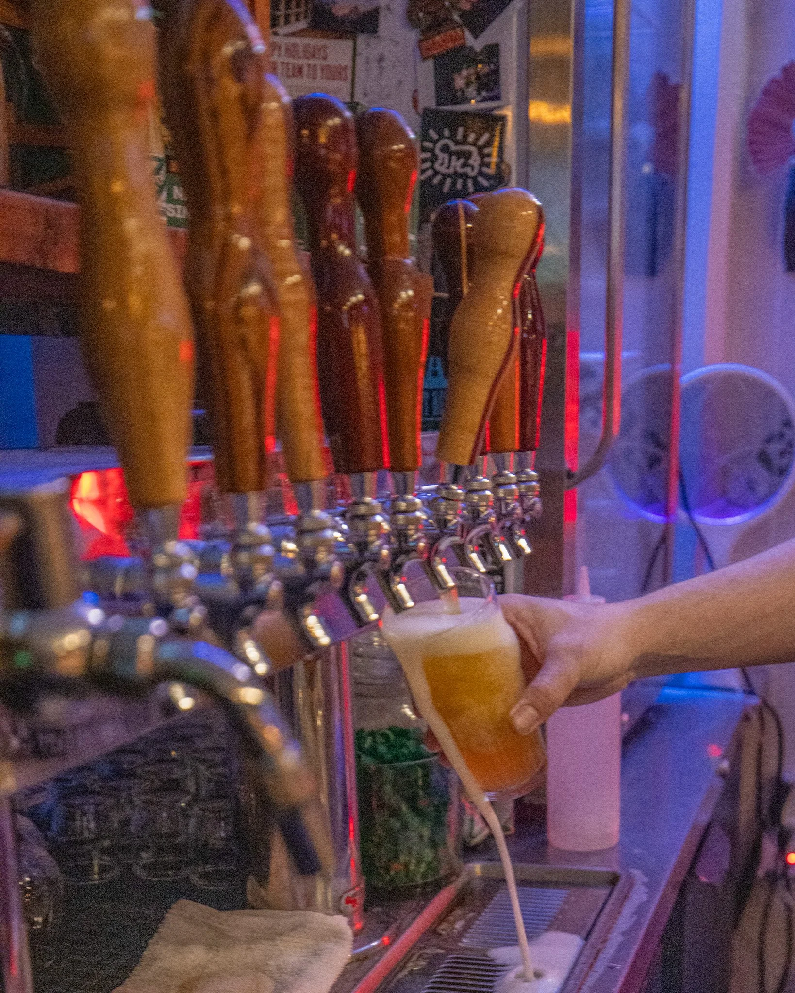 Person pouring a glass of beer from a tap at a bar or brewery with several beer taps visible.