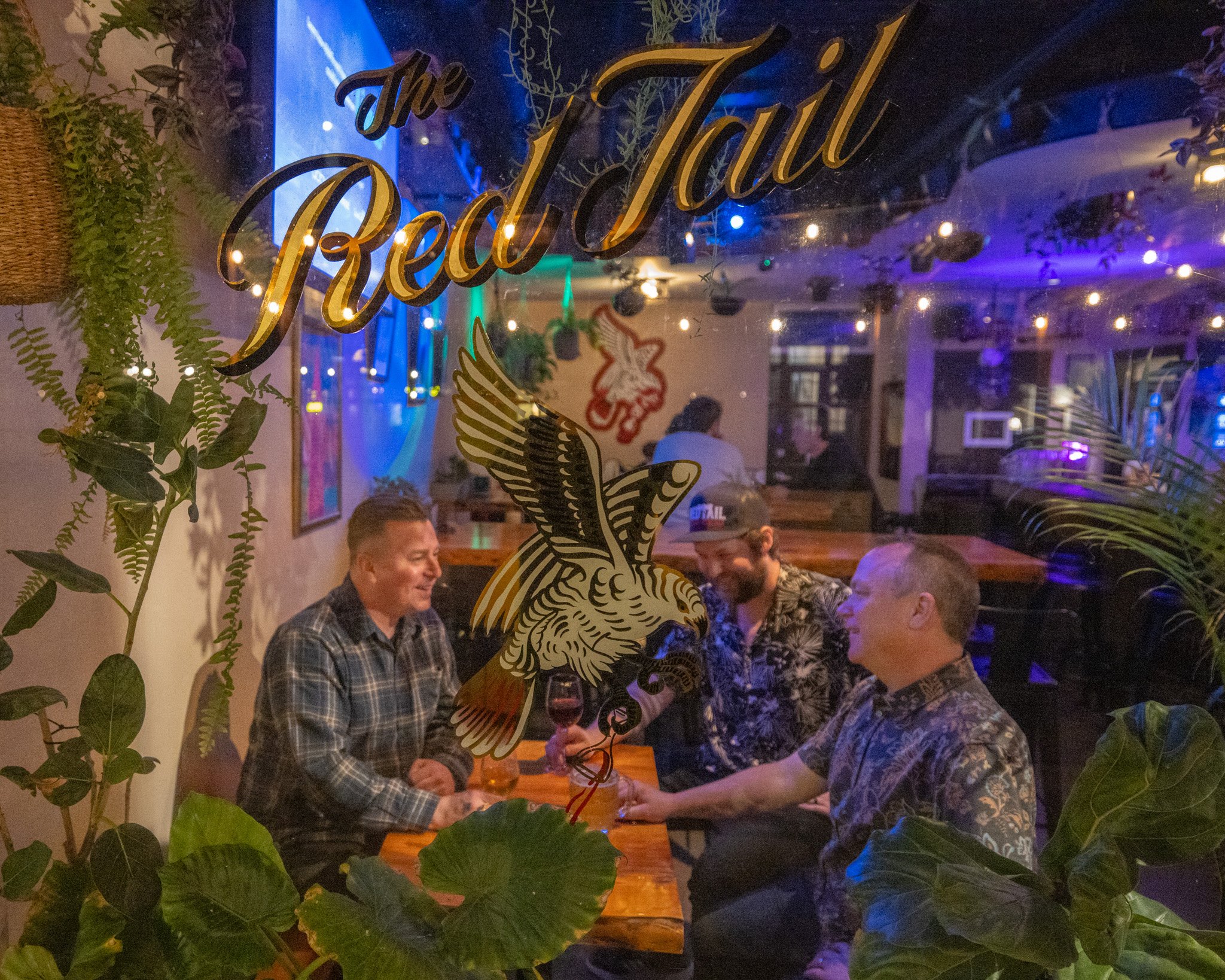 People socializing in a dimly lit restaurant or bar with a sign on the window that reads 'The Real'. The scene is decorated with string lights and plants, creating a cozy atmosphere.