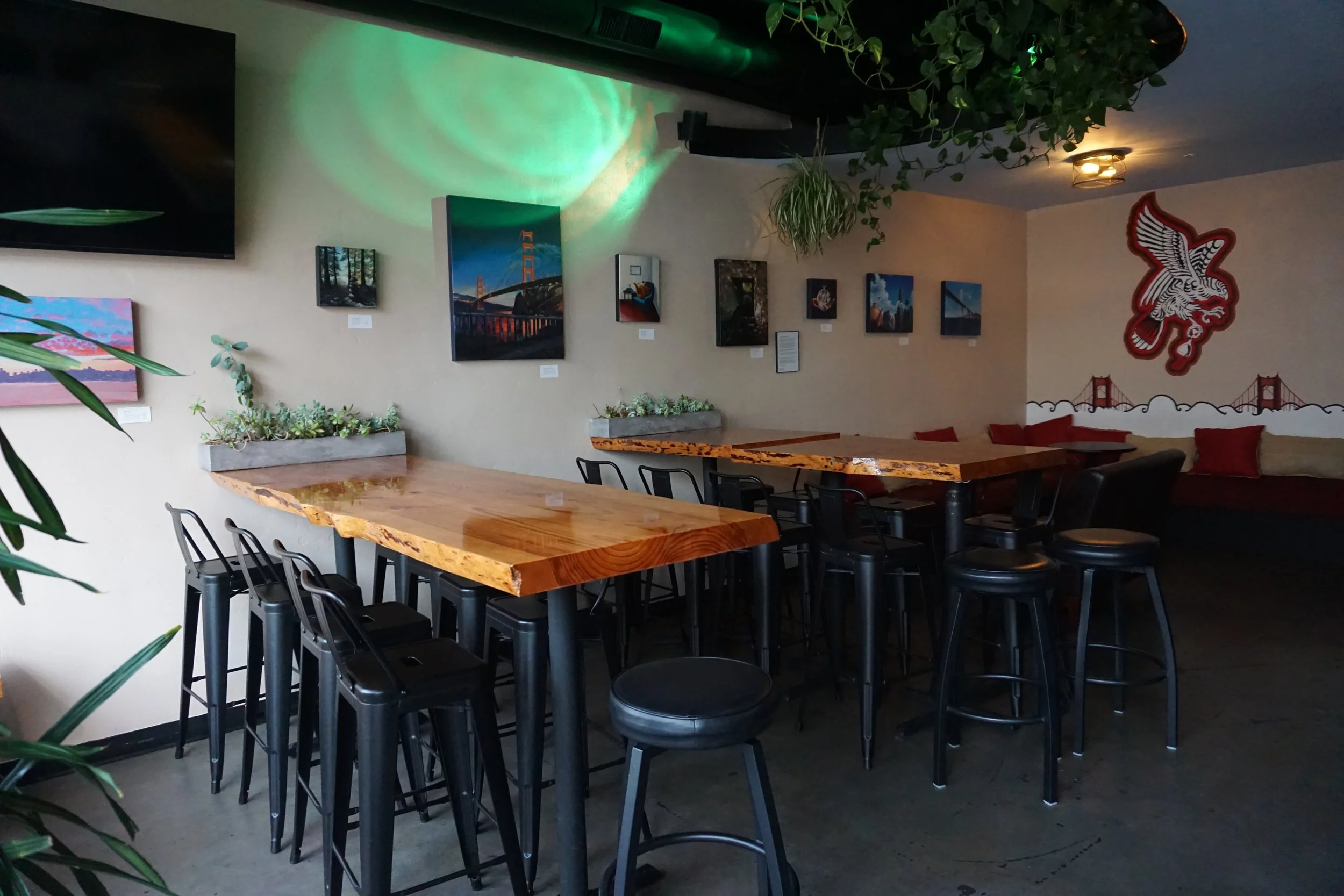 Interior of a modern cafe with wooden tables, black chairs, wall art, and greenery hanging from the ceiling. A large television is on the left wall, and a decorative red dragon sculpture with a cityscape mural is on the right wall.