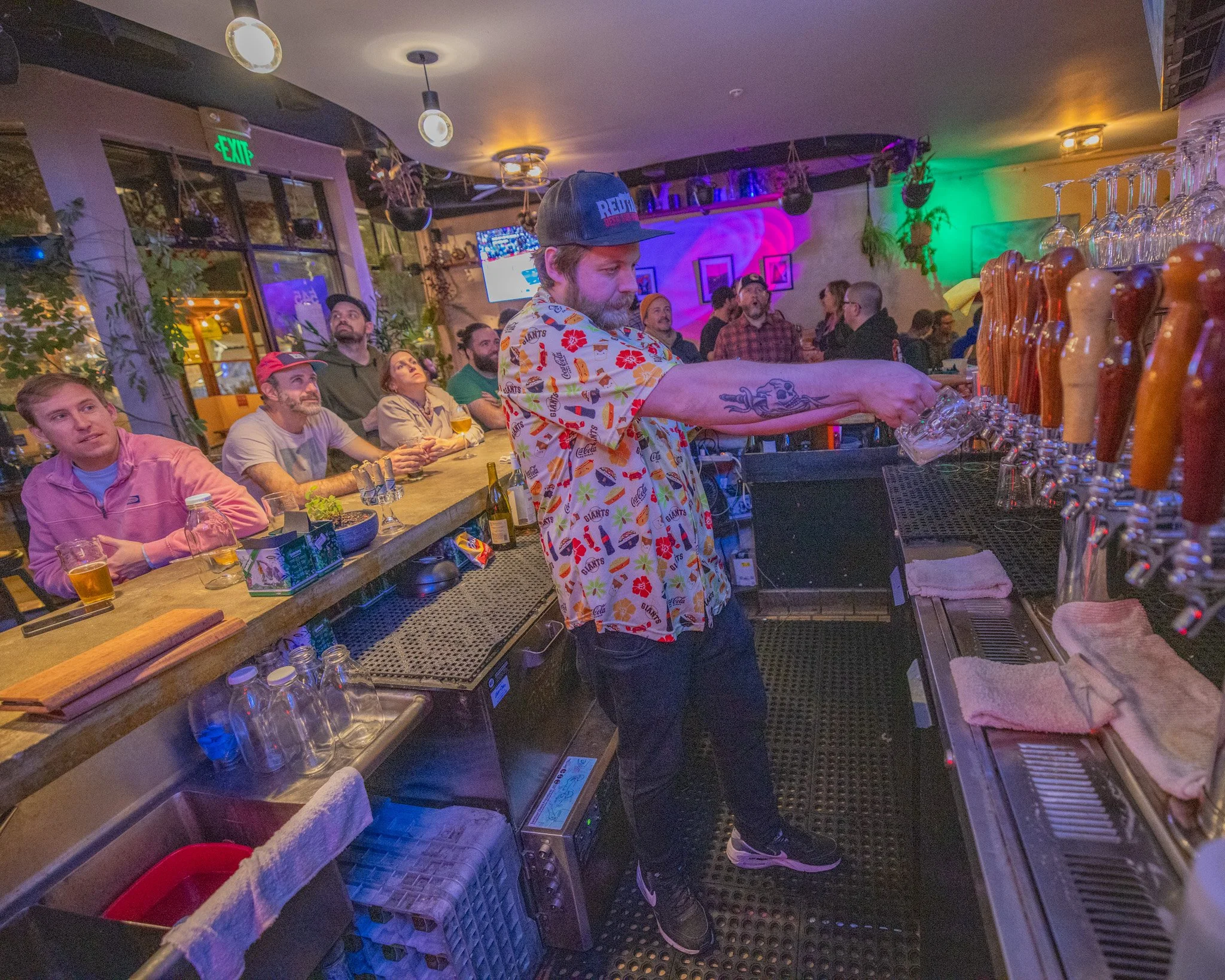  bartender serving drinks at a bar with a group of customers watching. The bar has glasses hanging upside down and colorful lighting.