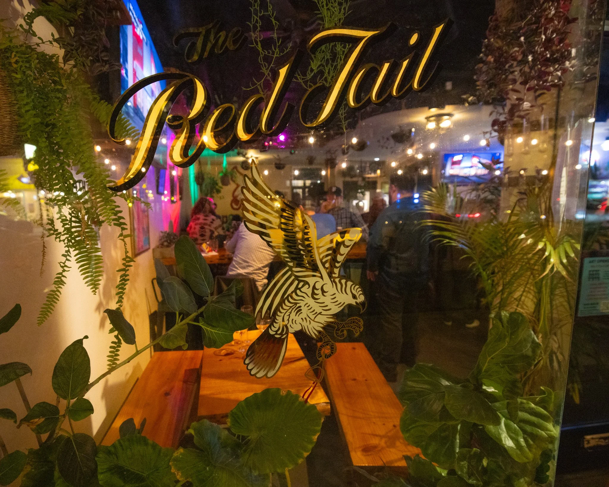 Inside view of a restaurant with neon lights, people dining, and a decorative decal of a bird and foliage on a glass door.