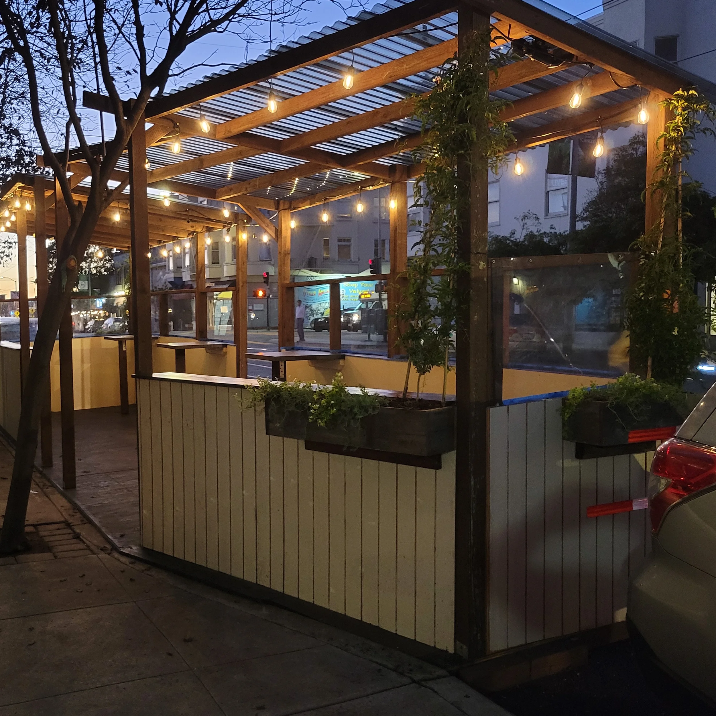 A wooden pergola with string lights hanging underneath, located on an urban street at dusk. There are trees and buildings in the background and a person walking by.