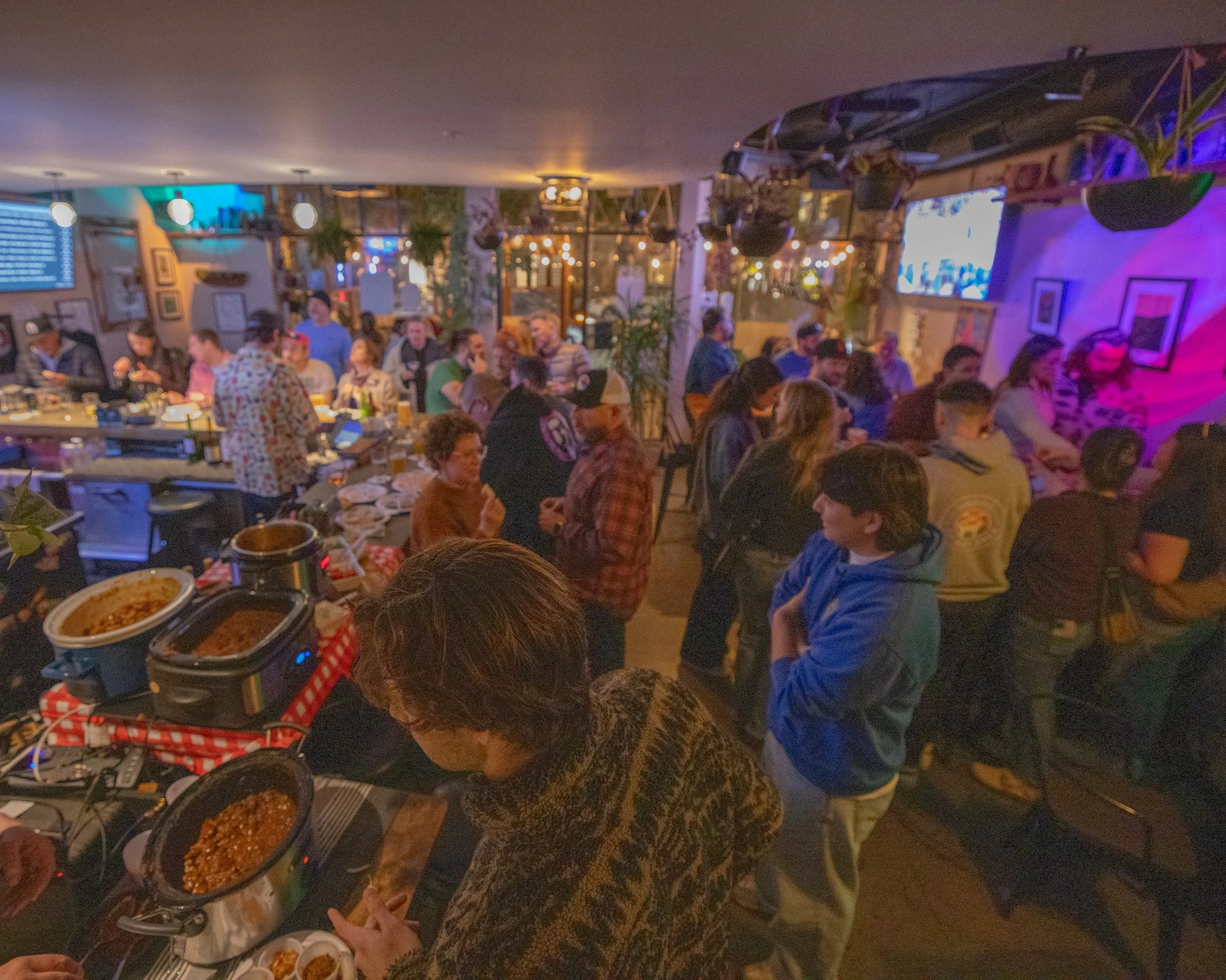 People gathered around a table at a restaurant enjoying and socializing, with food and drinks visible in the foreground, illuminated by warm ambient lighting.