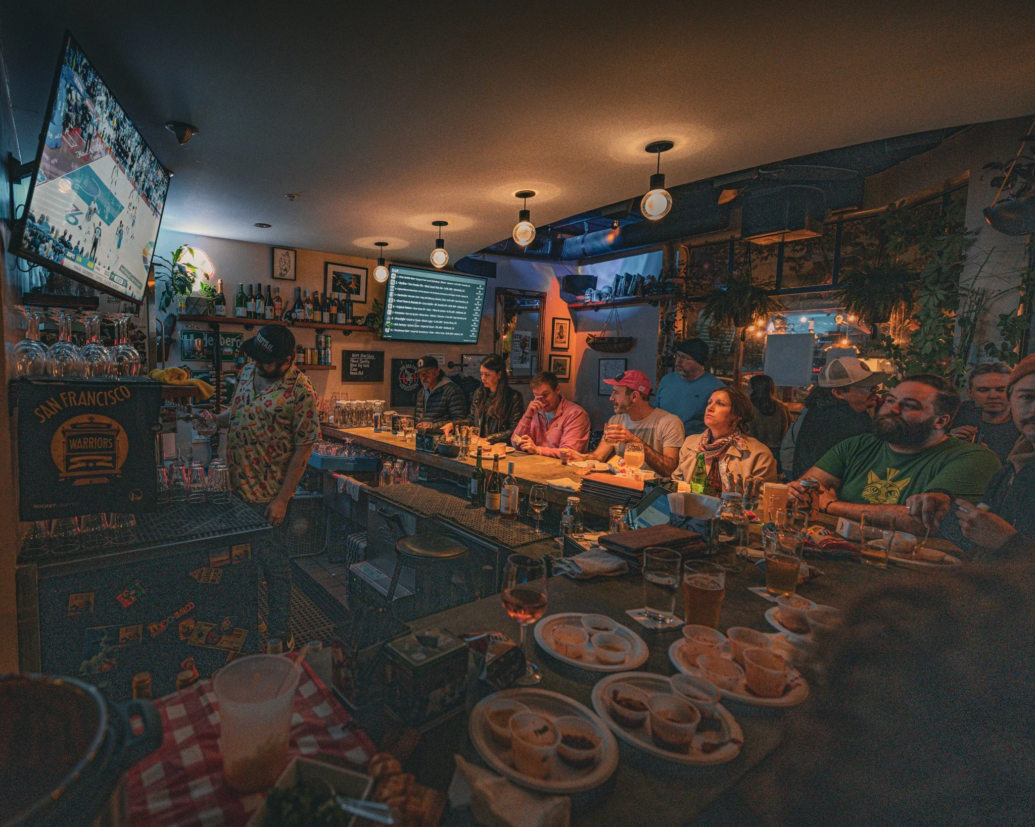 People dining and watching sports on TV in a cozy, dimly-lit bar or restaurant with drinks and food on the tables.