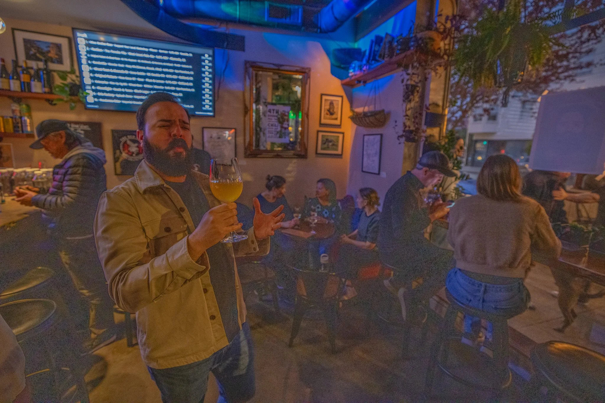 A man with a beard and wearing a tan jacket holding a wine glass in a dimly lit, busy restaurant or bar with multiple people, some wearing hats, and various framed pictures on the wall, illuminated with blue and warm lighting.