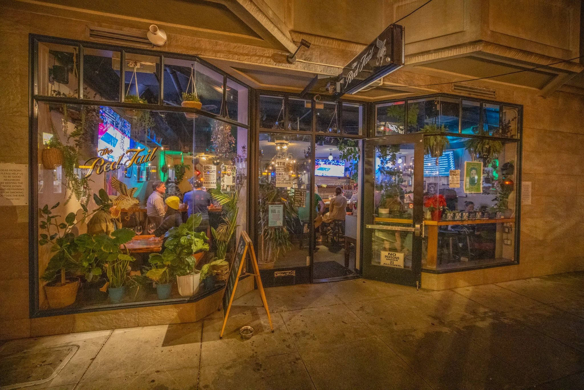 Nighttime view of a restaurant named 'The Red Tail' with glass walls, hanging plants, and potted plants outside, with people dining inside visible through the windows.