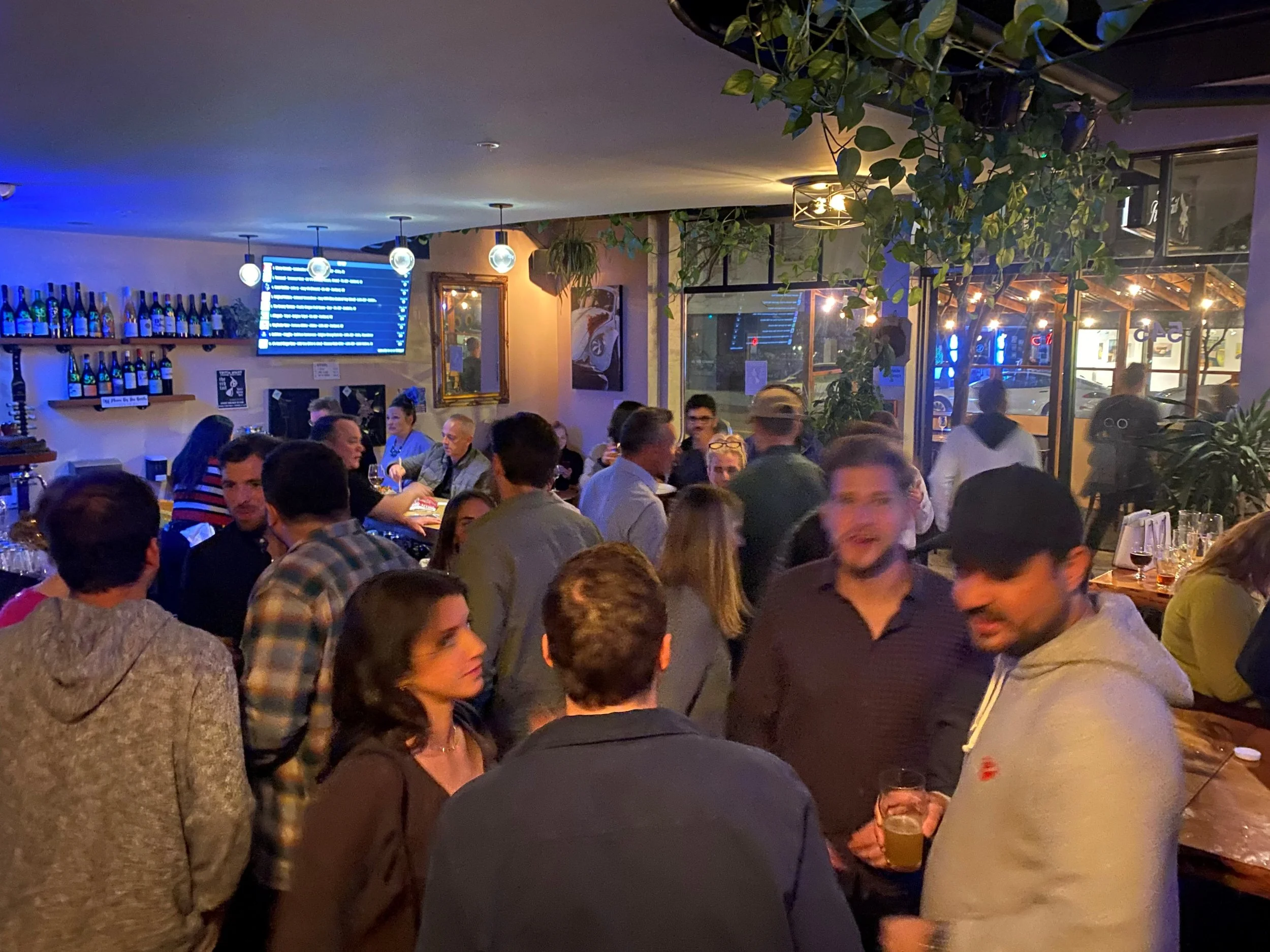 A crowded indoor restaurant or bar filled with people socializing. The space is decorated with plants hanging from the ceiling, and there is a display of bottles and a digital menu screen on the wall.