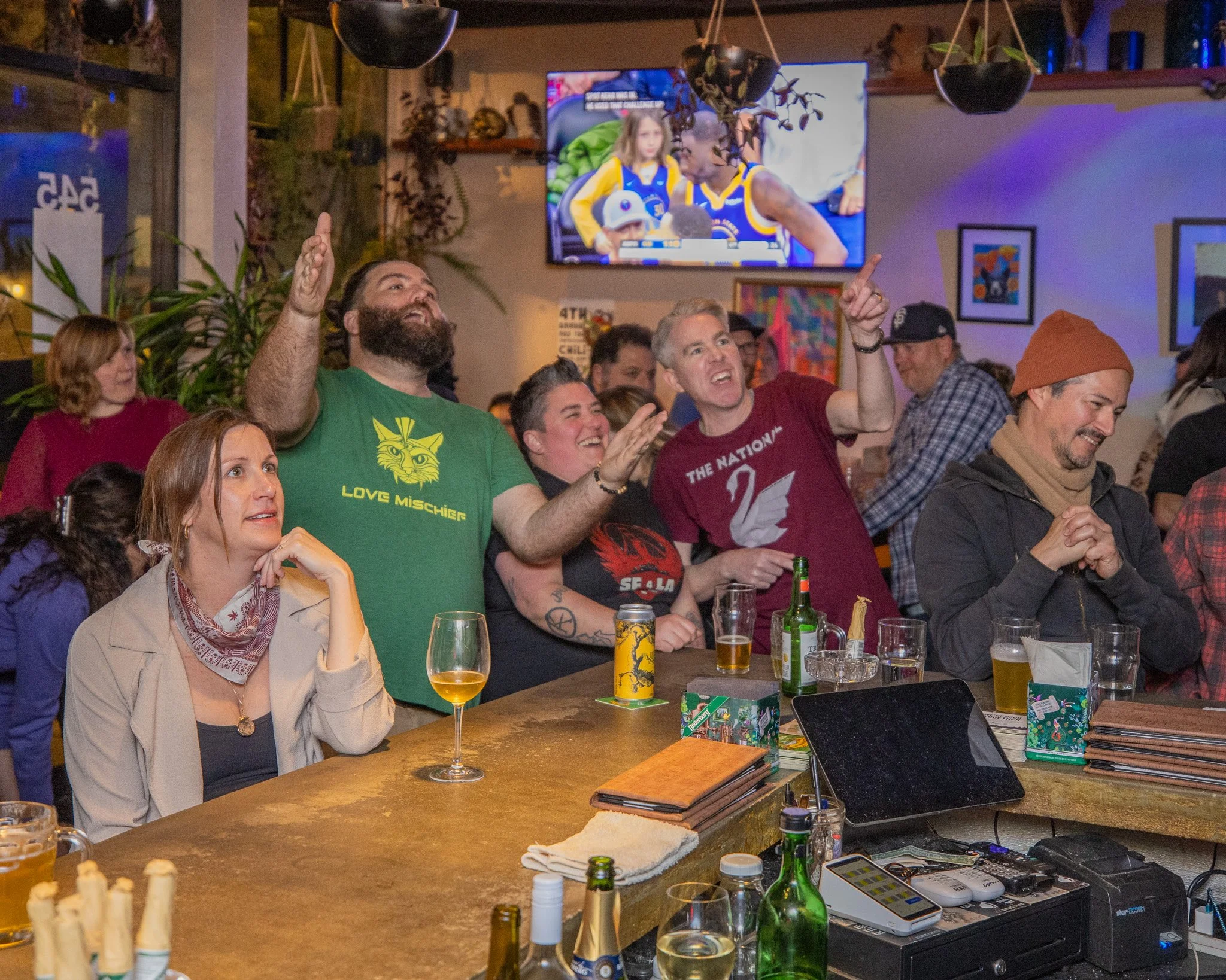 People gathered at a bar watching a basketball game on TV, cheering and enjoying drinks, with arcade games and various items on the bar counter.